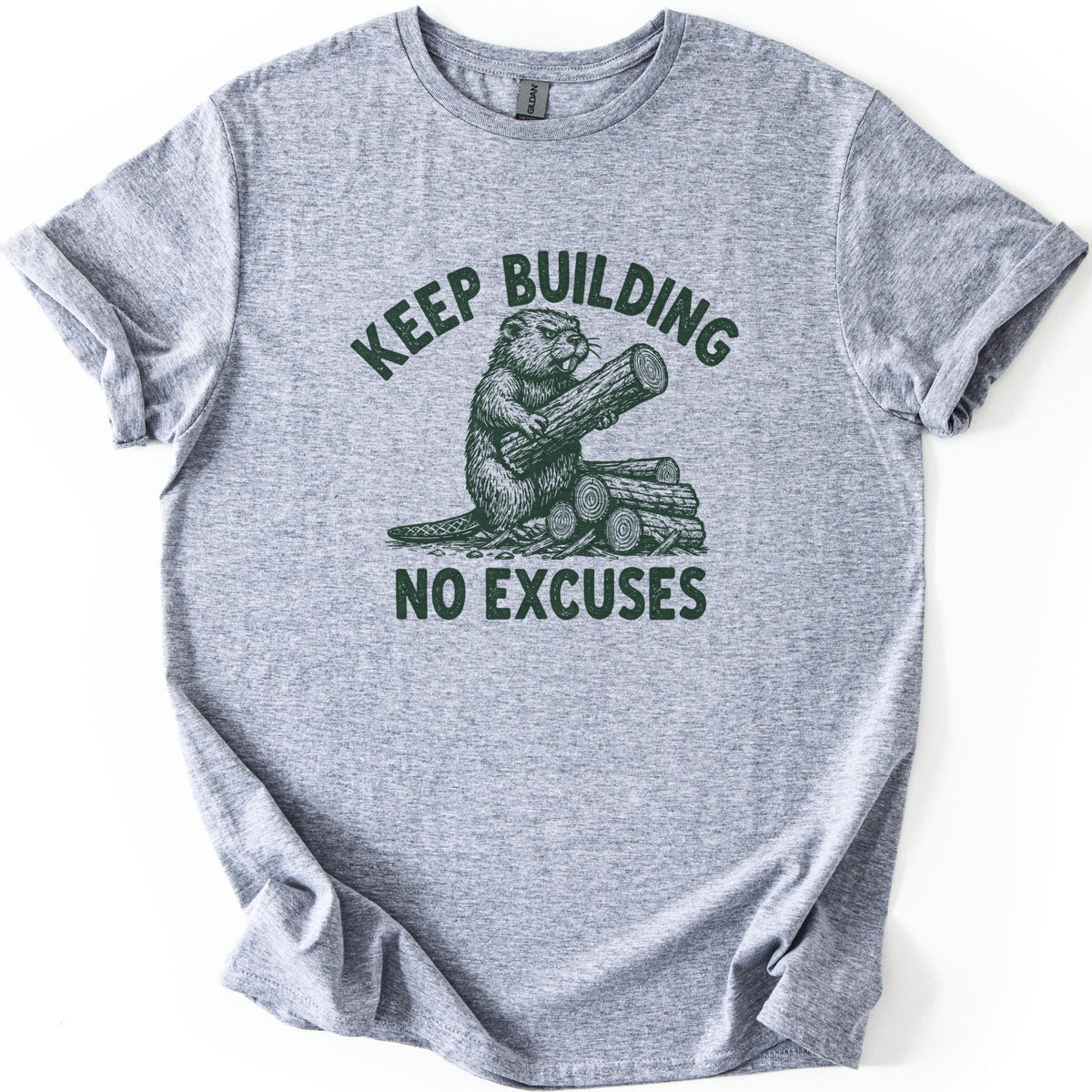 Keep Building Beaver Comfort Colors T-shirt in Sport Grey, laid flat. Dark green sketch-style artwork of a beaver hugging a log while sitting on a pile of cut logs, with Keep Building arched above and No Excuses below. Sleeves are rolled on a white background.