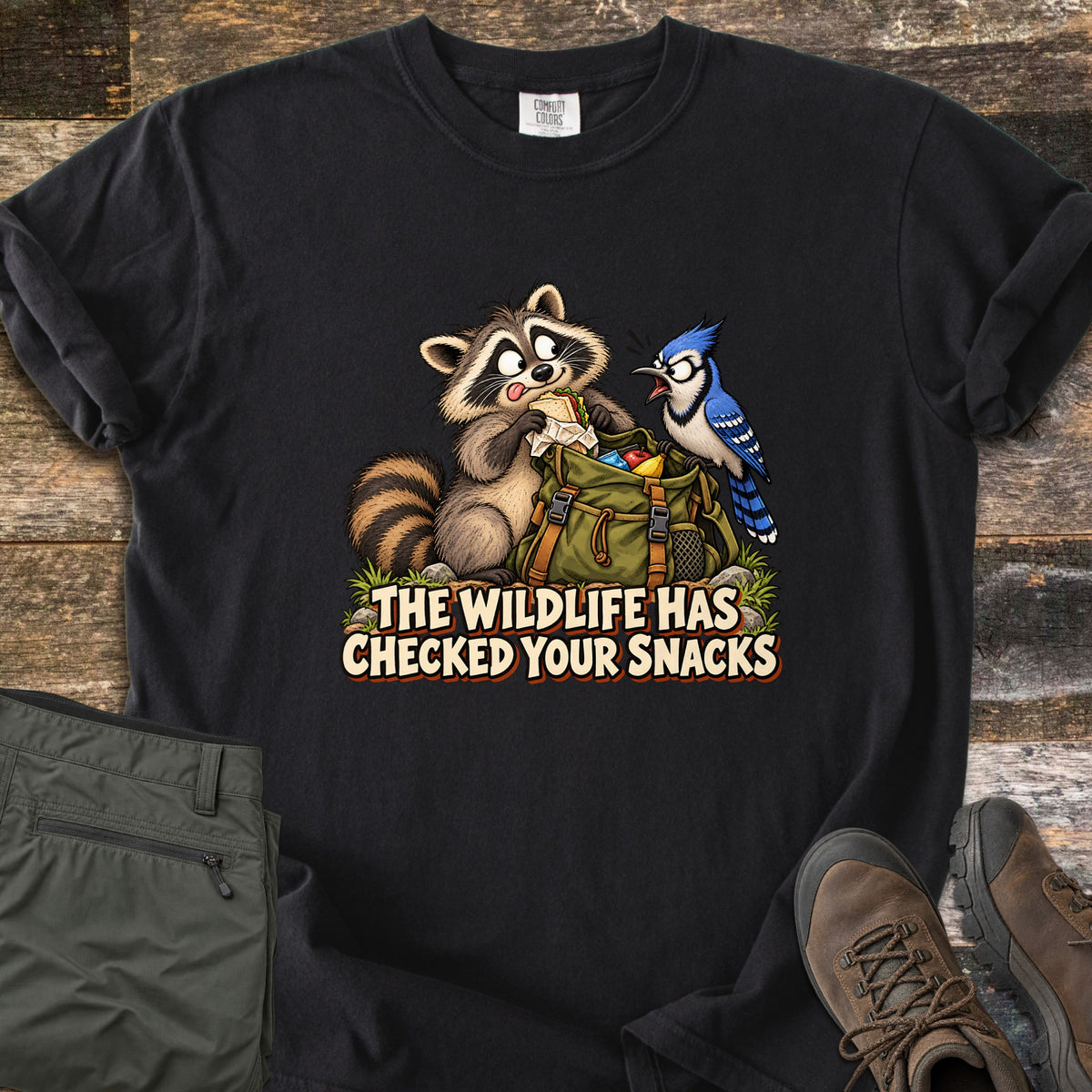 The Wildlife Has Checked Your Snacks Comfort Colors T-shirt in Black, laid flat. A cartoon raccoon eats a sandwich from a green backpack while a startled blue jay squawks at it, above the words The Wildlife Has Checked Your Snacks. Olive hiking pants and brown boots lie on weathered wood.