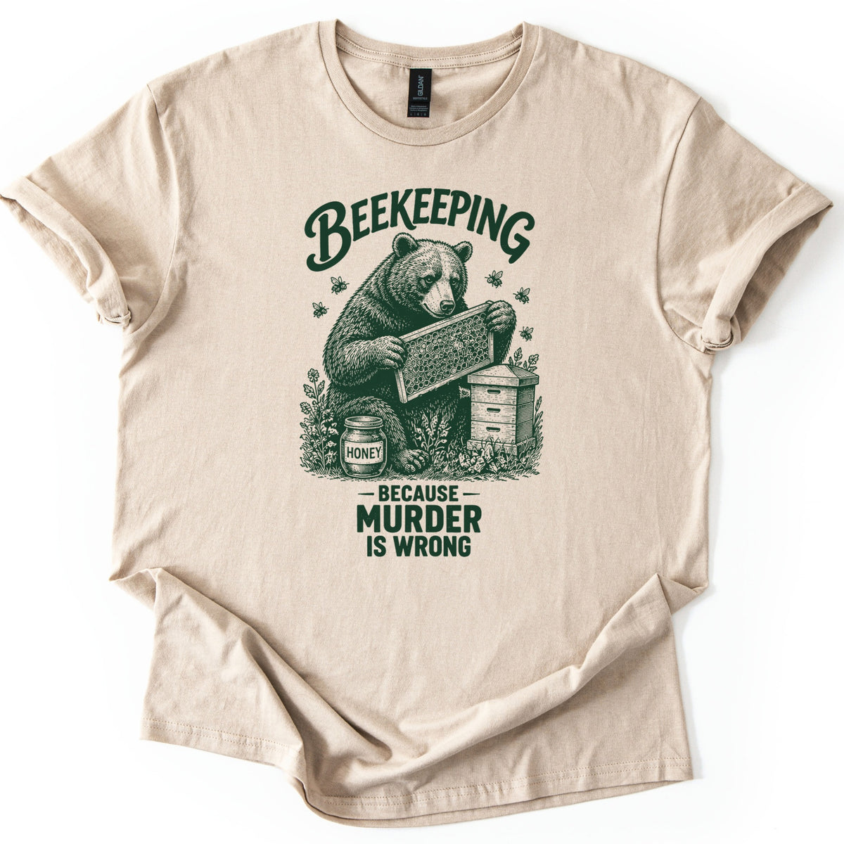 Beekeeping Murder Bear Comfort Colors T-shirt in Sand, laid flat. A dark green etched drawing of a bear holding a honeycomb frame by a beehive and a jar labeled Honey, with bees flying, under the word Beekeeping and above Because Murder Is Wrong. Shown on white with cuffed sleeves.