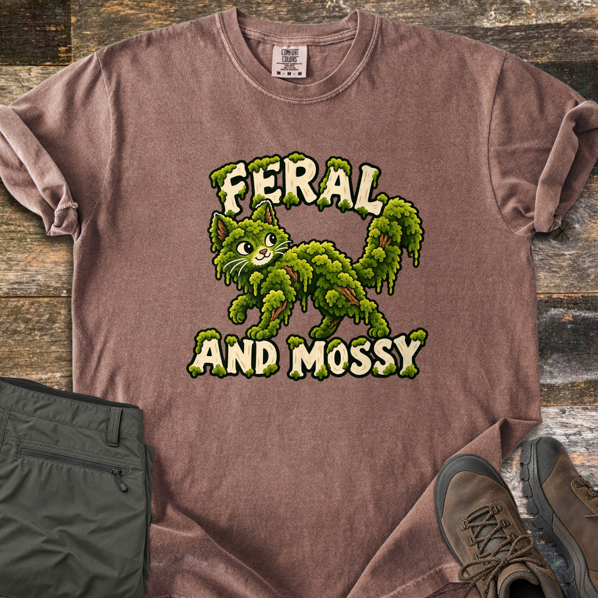 Feral and Mossy Comfort Colors T-shirt in Espresso, laid flat. A cartoon cat made of green moss with bits of bark walks between the words Feral and Mossy in cream letters dripping with moss. Folded olive pants and brown hiking boots sit on a weathered wood floor.