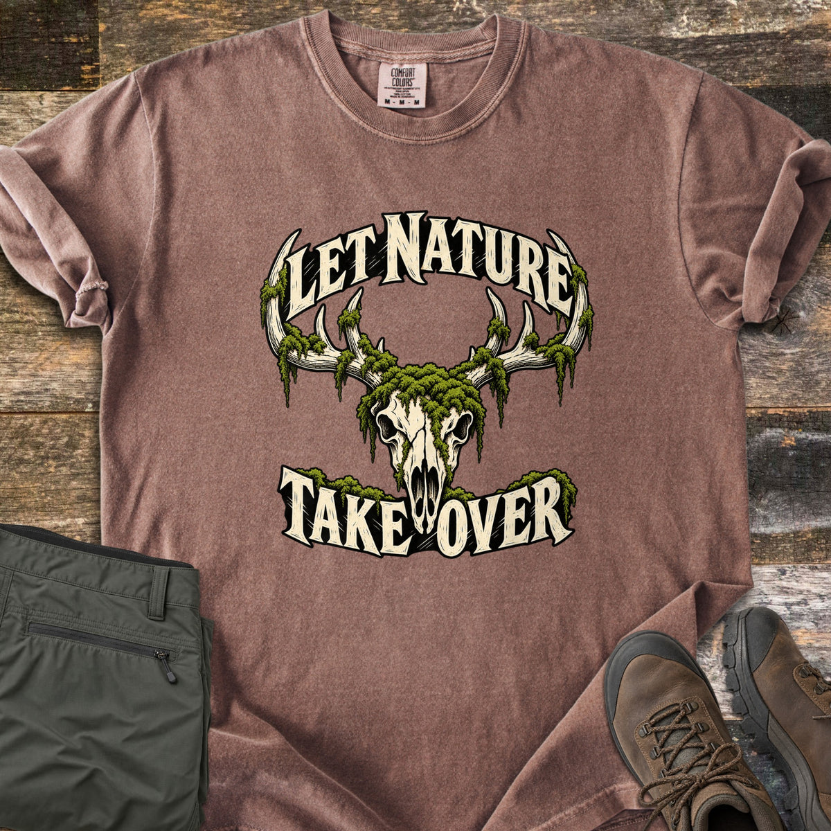 Let Nature Take Over Comfort Colors T-shirt in Espresso, laid flat. A moss-covered deer skull with antlers sits between the words Let Nature Take Over in cream lettering. Folded olive hiking pants and brown hiking boots lie beside it on weathered wood planks.