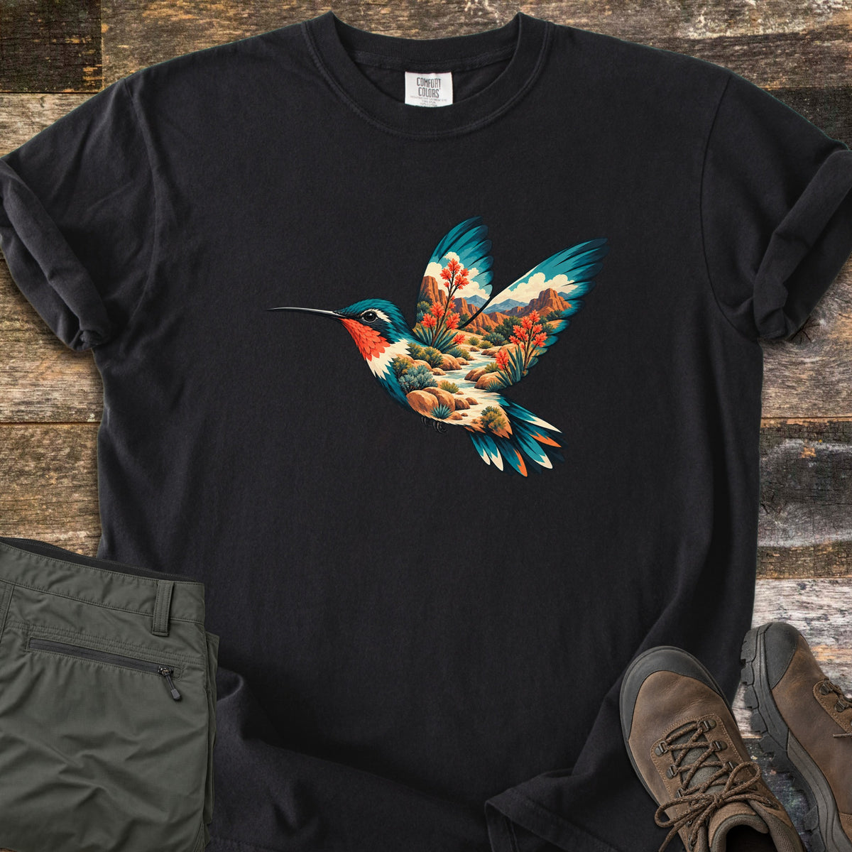 Hummingbird Canyon Comfort Colors T-shirt in Black, laid flat. A hummingbird in flight whose body and wings are filled with a desert canyon scene of red rock cliffs, a winding stream, boulders and red-blooming plants. Olive hiking pants and brown boots sit on weathered wood.