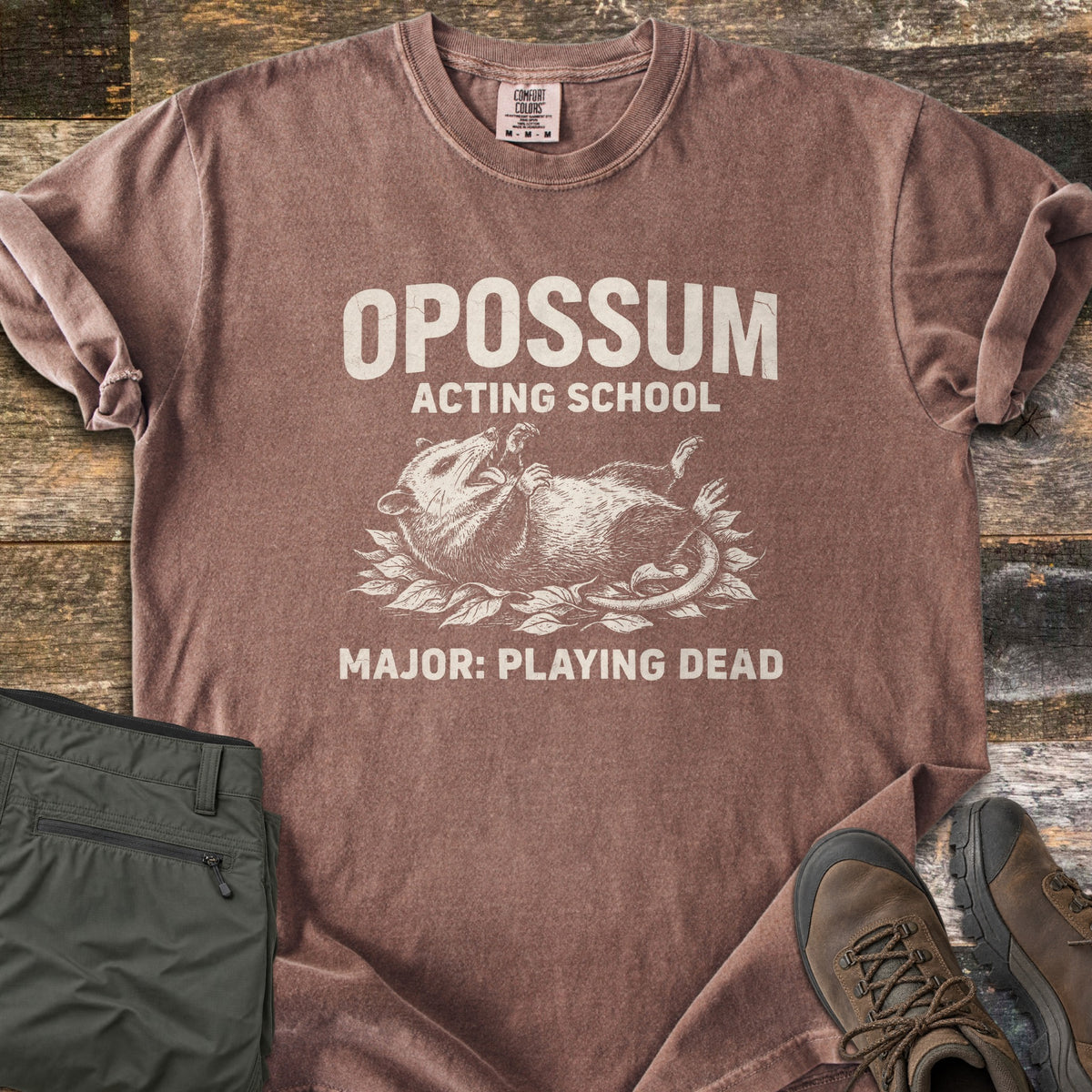 Opossum Acting School Comfort Colors T-shirt in Espresso, laid flat. An etched drawing of an opossum lying on its back on leaves, mouth open, with the words Opossum Acting School above and Major: Playing Dead below. Sleeves rolled on wood planks beside green pants and brown hiking boots.