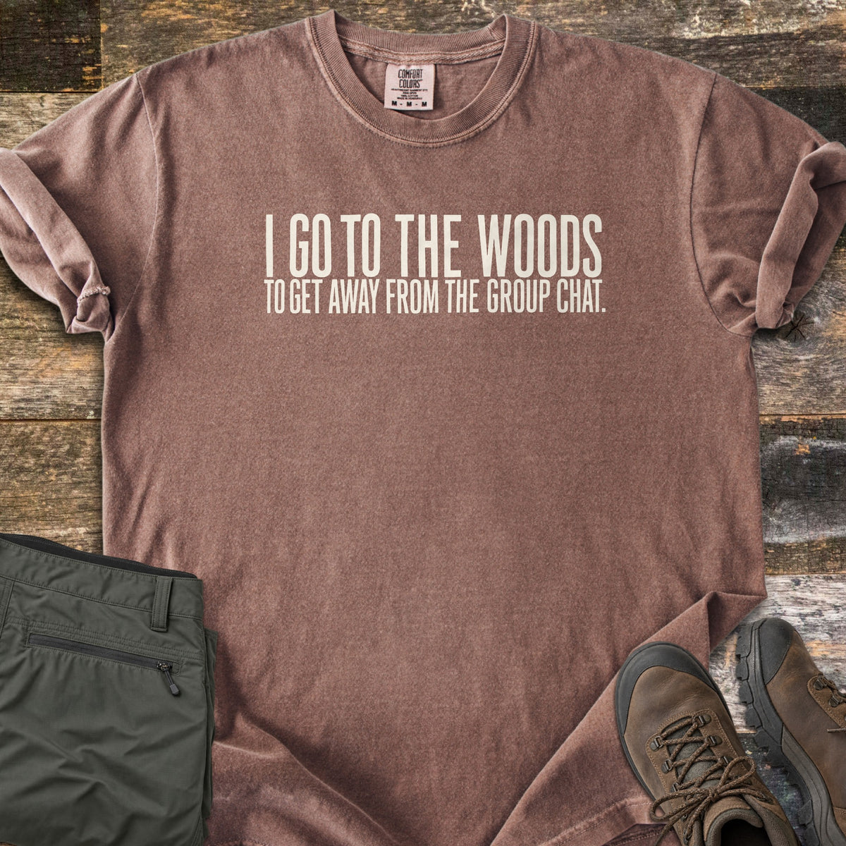 Away From The Group Chat Comfort Colors T-shirt in Espresso, laid flat. Off-white block letters read I GO TO THE WOODS above TO GET AWAY FROM THE GROUP CHAT. The shirt has rolled sleeves and lies on weathered wood boards beside folded olive green pants and brown hiking boots.