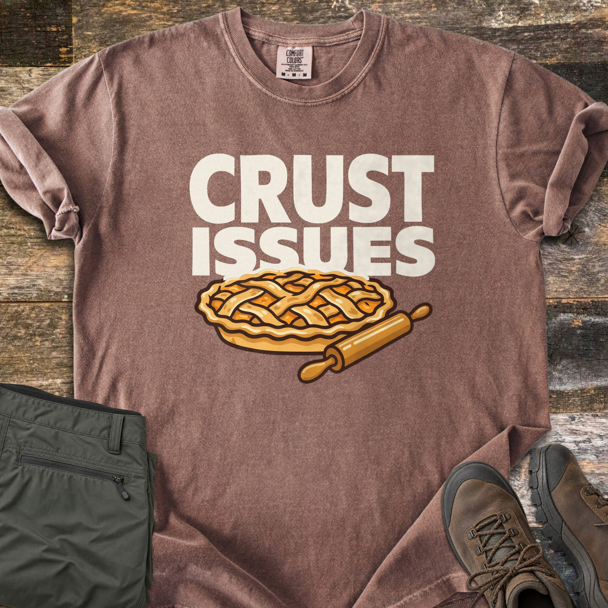Crust Issues Comfort Colors T-shirt in Espresso, laid flat. The words CRUST ISSUES in off-white block letters sit above a cartoon lattice-top pie and a wooden rolling pin. Olive pants and brown hiking boots lie beside it on a rustic wood surface.