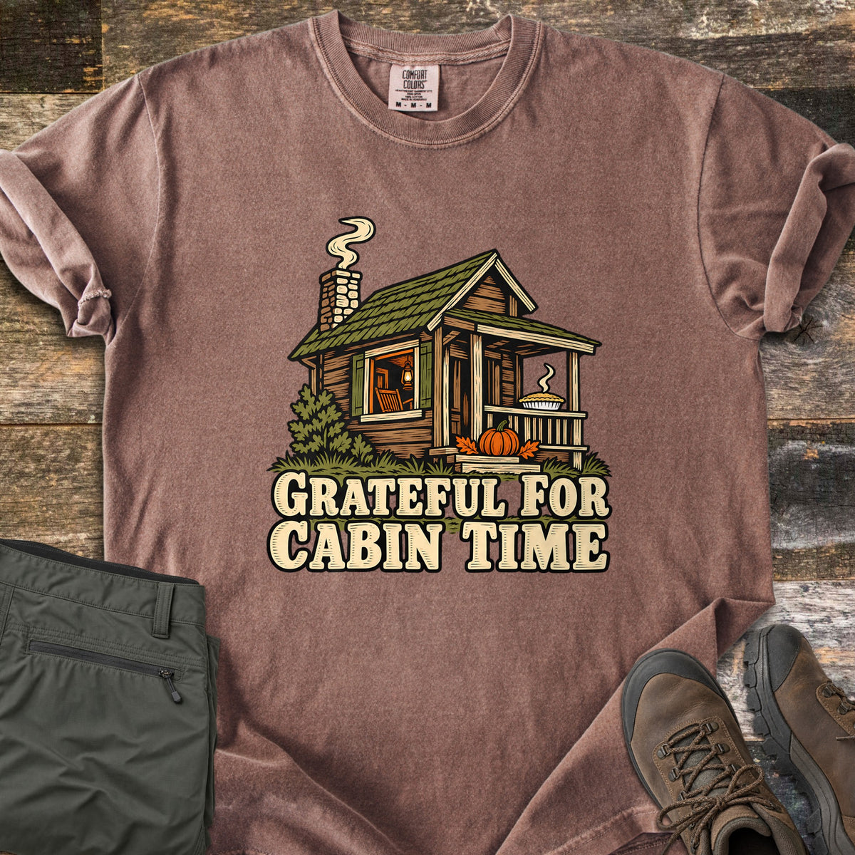 Grateful For Cabin Time Comfort Colors T-shirt in Espresso, laid flat. Print of a log cabin with a green roof, smoking chimney, a pie on the porch rail and a pumpkin on the steps, above the words Grateful For Cabin Time. Green pants and brown hiking boots lie on weathered wood.