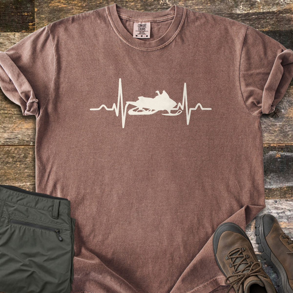 Snowmobile Heartbeat Comfort Colors T-shirt in Espresso, laid flat. A cream heartbeat line runs across the chest with a snowmobile silhouette in its center, with no words. Sleeves are rolled, on weathered wood planks beside folded olive pants and brown hiking boots.