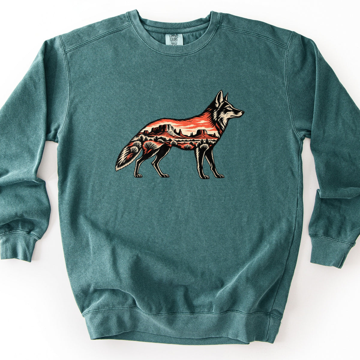 Desert Mesa Coyote Comfort Colors crewneck sweatshirt in Blue Spruce, laid flat. A coyote in side profile, its body filled with a desert scene of red mesas, orange sky and sagebrush, in black, cream and red-orange tones, with no words. The sweatshirt lies on a white background.