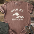 Be Superior Comfort Colors T-shirt in Espresso, laid flat. Cream text reads Don't Be Salty, arched over a Lake Superior silhouette topped with a small crown, with Be Superior below. Sleeves rolled, on weathered wood with olive hiking pants and brown hiking boots.