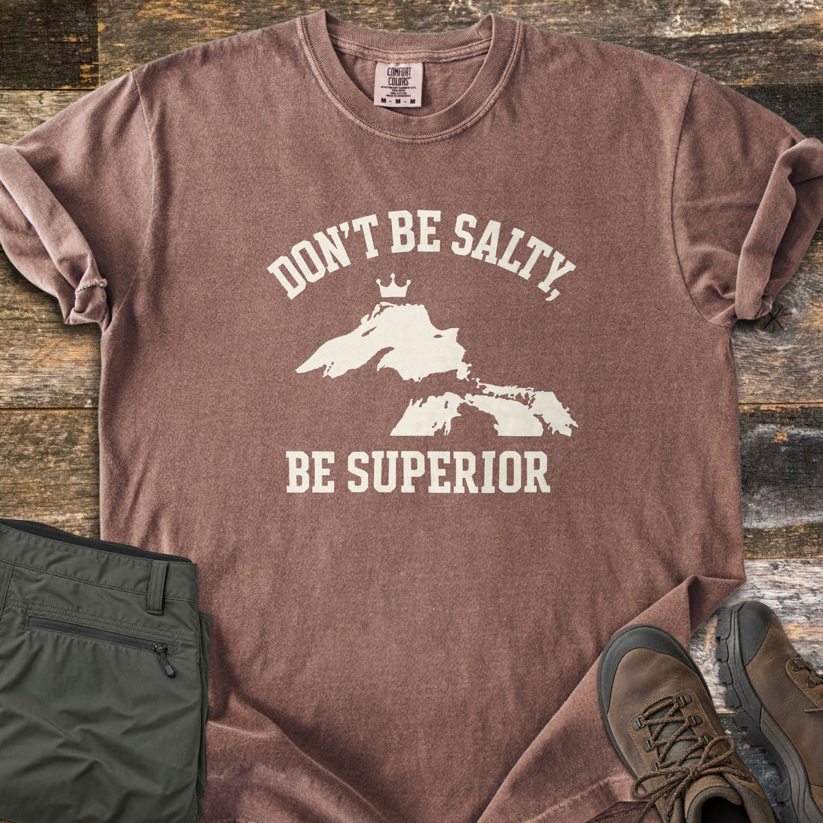 Be Superior Comfort Colors T-shirt in Espresso, laid flat. Cream text reads Don't Be Salty, arched over a Lake Superior silhouette topped with a small crown, with Be Superior below. Sleeves rolled, on weathered wood with olive hiking pants and brown hiking boots.