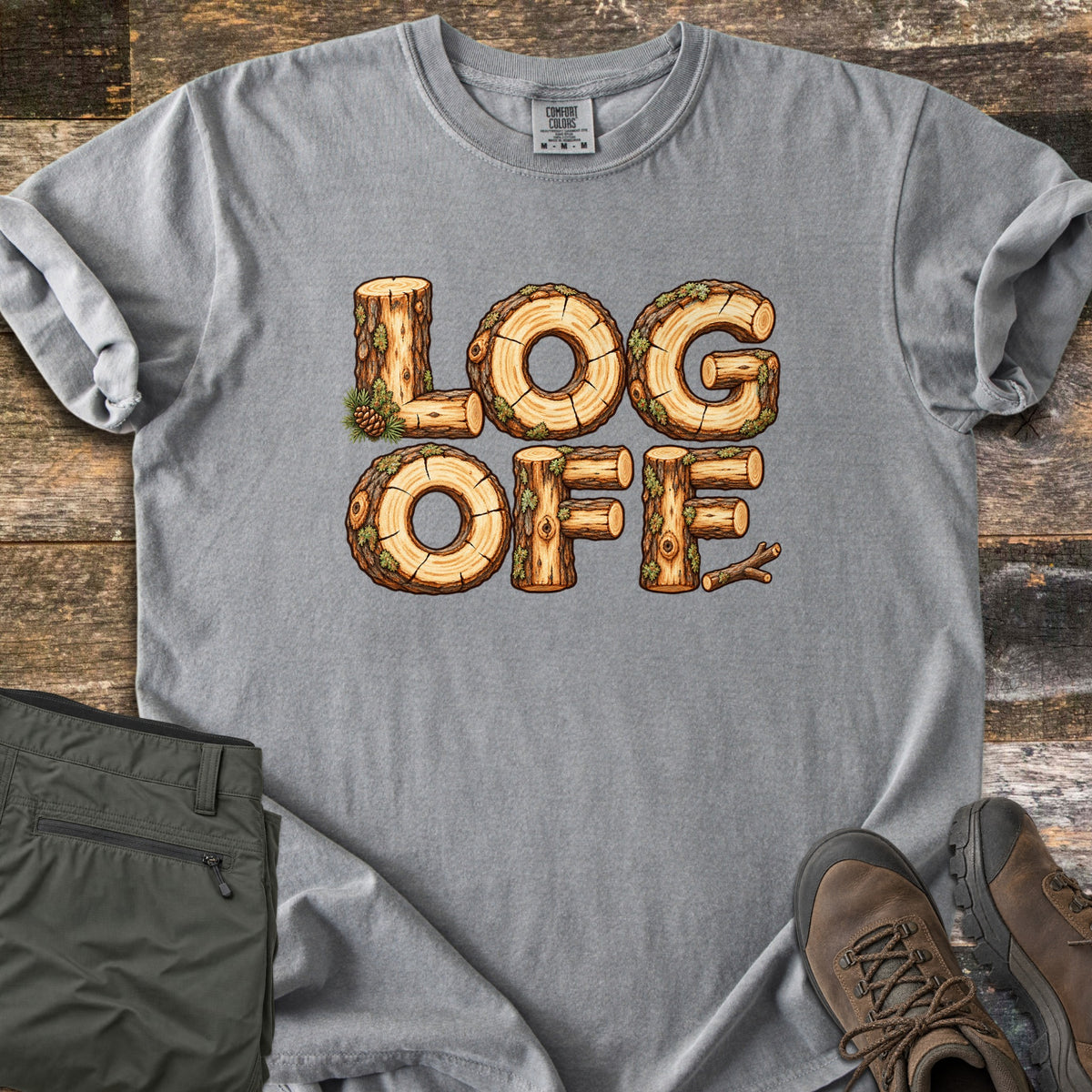 Log Off Comfort Colors T-shirt in Grey, laid flat. The words Log Off are spelled in letters made of cut wooden logs with bark, moss and a pine cone. The shirt lies on weathered wood planks beside folded olive pants and brown hiking boots.