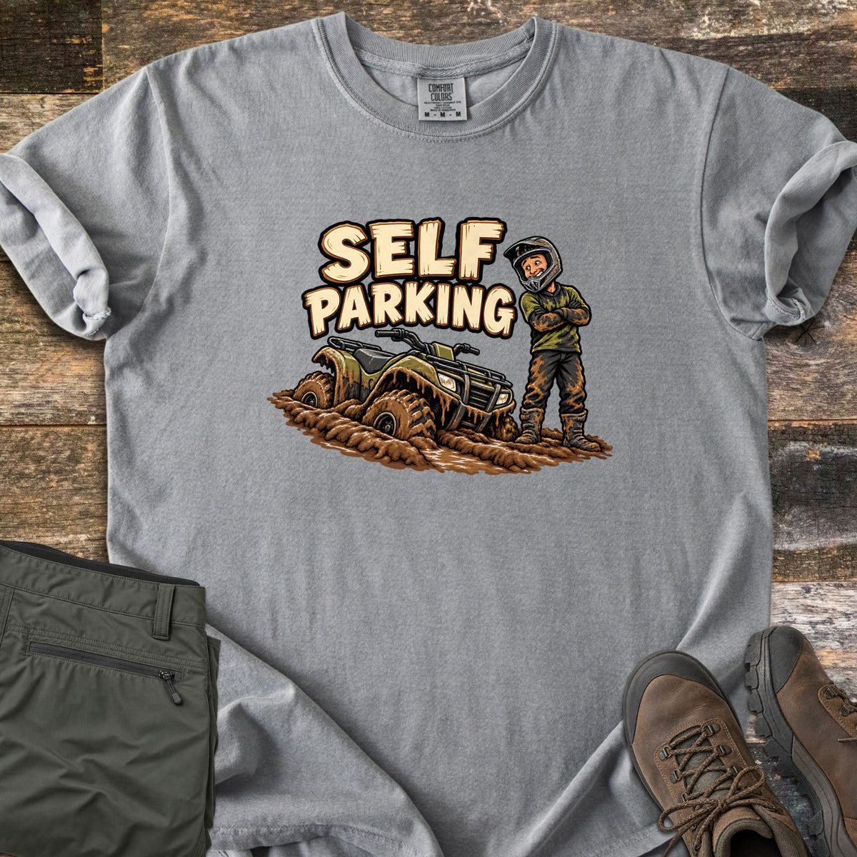 Self Parking Comfort Colors T-shirt in Grey, laid flat. A cartoon rider in a helmet stands arms crossed, covered in mud, beside an ATV sunk in mud under the words Self Parking. Folded olive pants and brown hiking boots sit on a wood plank background.