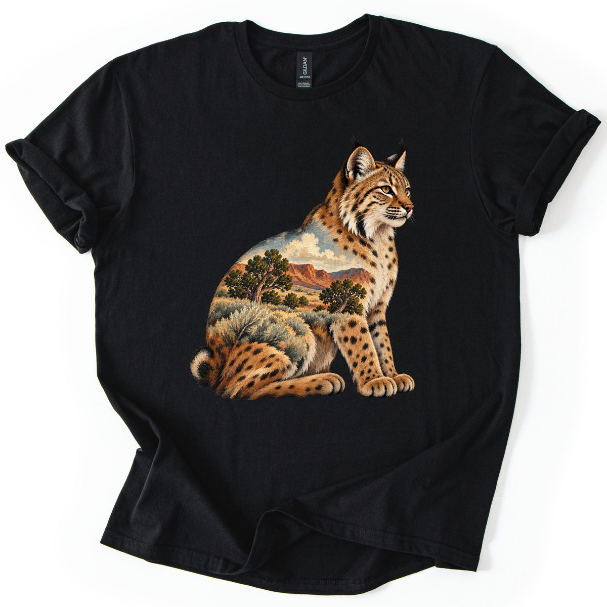 Bobcat Sagebrush Canyon Comfort Colors T-shirt in Black, laid flat. A painted bobcat sits facing right, its body filled with a desert scene of red canyon cliffs, green trees, sagebrush and clouds. No words are printed. The shirt lies on a white surface with cuffed sleeves.