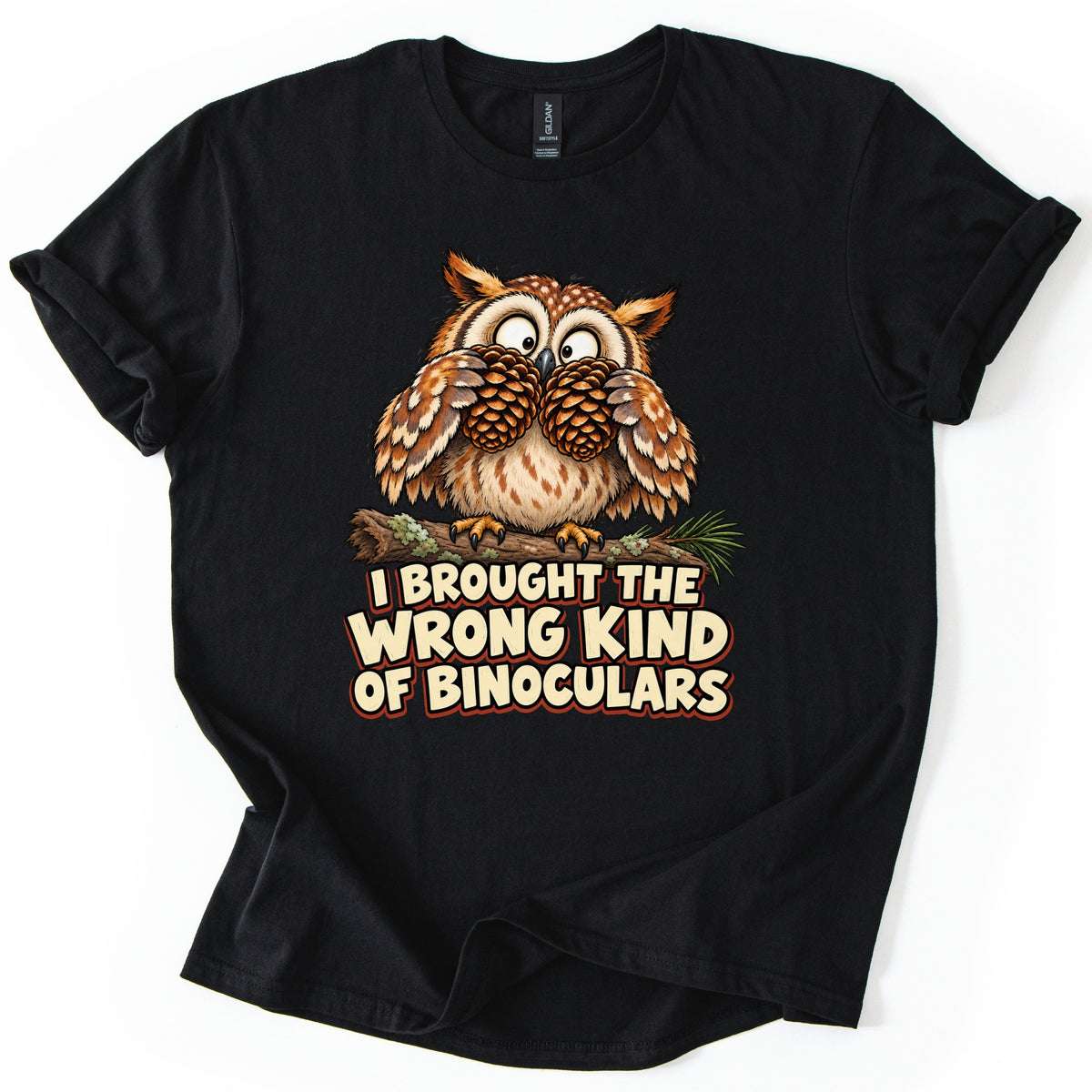 I Brought The Wrong Kind Of Binoculars Comfort Colors T-shirt in Black, laid flat. A wide-eyed cartoon owl perched on a mossy branch holds two pine cones up to its eyes like binoculars, above the words I Brought The Wrong Kind Of Binoculars in cream letters. Rolled sleeves, on a white background.