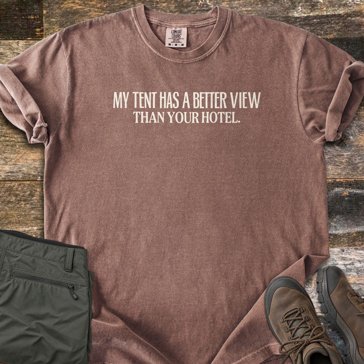 Better View Than Your Hotel Comfort Colors T-shirt in Espresso, laid flat. Cream text reads My Tent Has a Better View Than Your Hotel. Sleeves are cuffed, on a weathered wood surface with folded olive hiking pants and brown hiking boots.