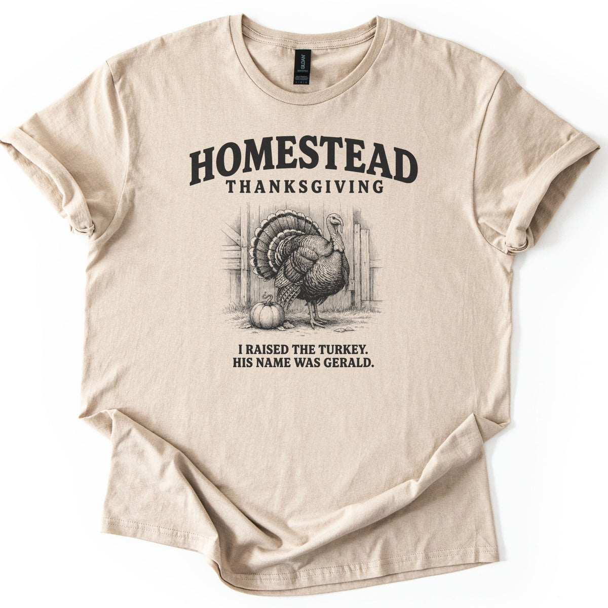 Homestead Thanksgiving Comfort Colors T-shirt in Sand, laid flat. Black ink sketch of a fanned-tail turkey beside a pumpkin by a wooden barn, under arched Homestead Thanksgiving and above I Raised the Turkey. His Name Was Gerald. Rolled sleeves, white background.
