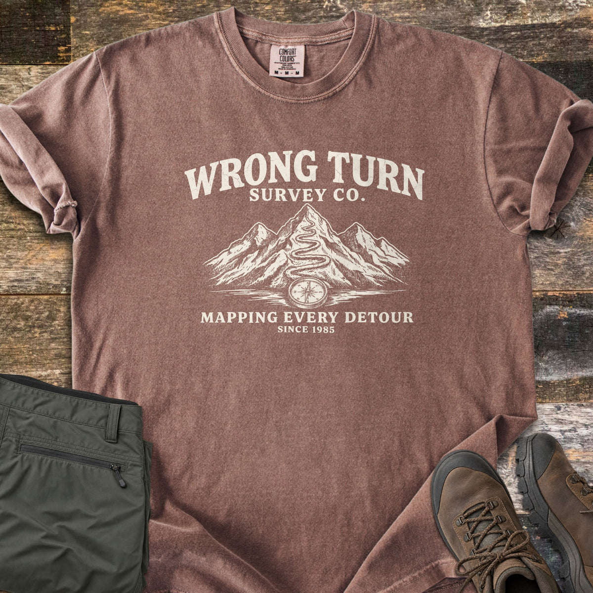 Wrong Turn Survey Co Comfort Colors T-shirt in Espresso, laid flat. Cream print of the words Wrong Turn Survey Co. above snowy mountains with a winding trail leading to a compass, then Mapping Every Detour, Since 1985. Shown on wood boards with olive pants and brown hiking boots.