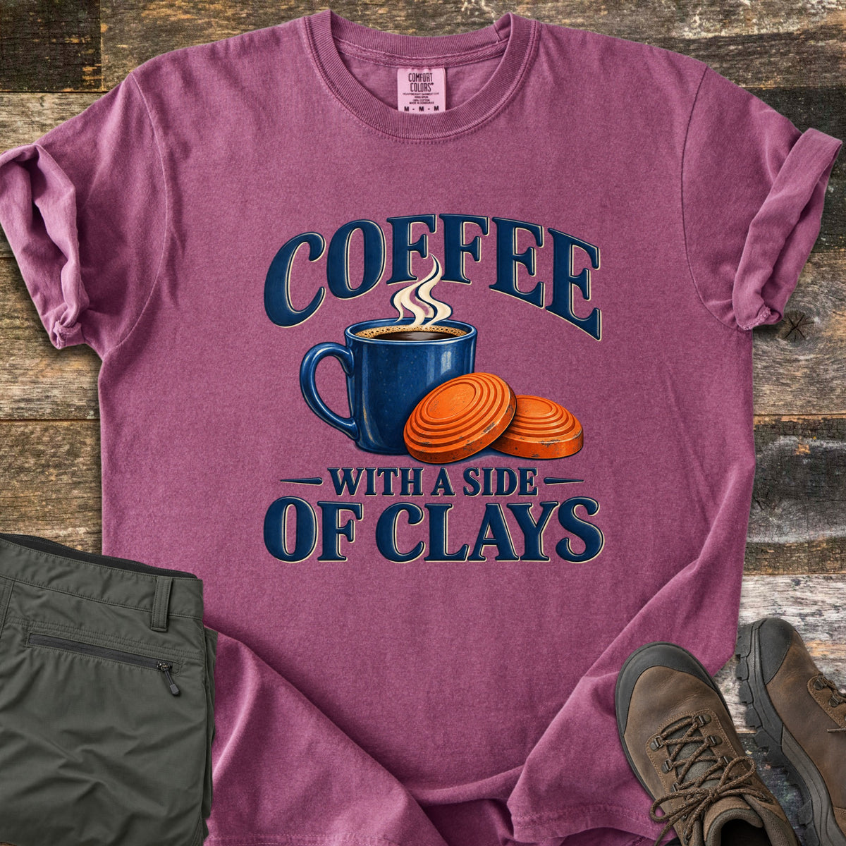 Coffee with a Side of Clays Comfort Colors T-shirt in Berry, laid flat. A steaming blue mug of coffee beside two orange clay targets, with the words Coffee with a Side of Clays in navy. Sleeves rolled, on rustic wood with folded green pants and brown hiking boots.