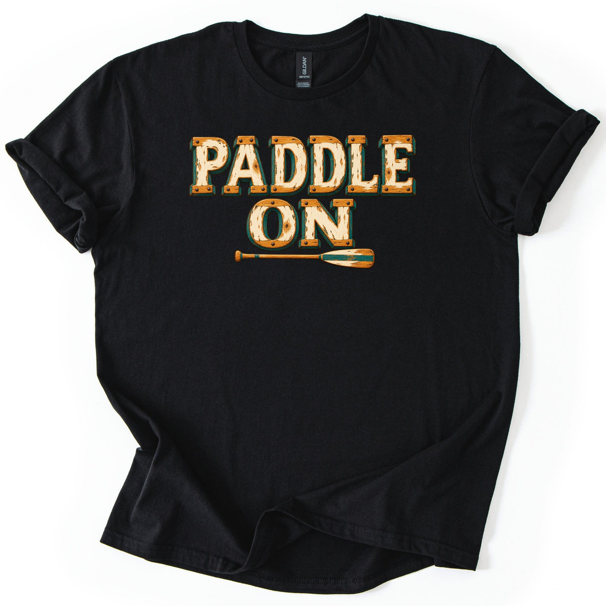 Paddle On Comfort Colors T-shirt in Black, laid flat. PADDLE ON is printed in distressed cream letters framed like riveted orange wood planks with a green outline, above a wooden paddle with a green-striped blade. Sleeves rolled, on a white surface.