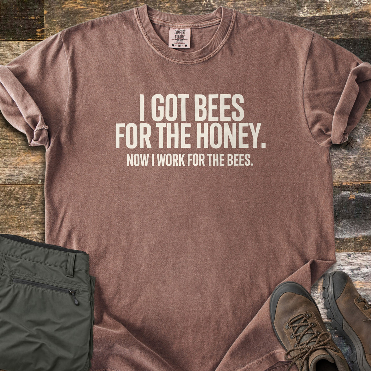 Work For The Bees Comfort Colors T-shirt in Espresso, laid flat. Cream block letters read I GOT BEES FOR THE HONEY. NOW I WORK FOR THE BEES. The shirt lies with cuffed sleeves on weathered wood planks beside folded olive green pants and brown hiking boots.