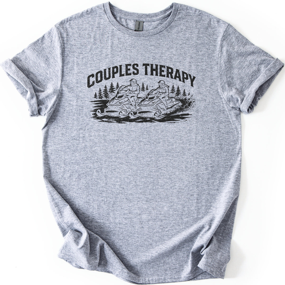 Couples Therapy Snowmobile Comfort Colors T-shirt in Sport Grey, laid flat. Black print of the arched words Couples Therapy above two helmeted riders on snowmobiles racing side by side through snow with pine trees behind. Sleeves are cuffed on a white background.