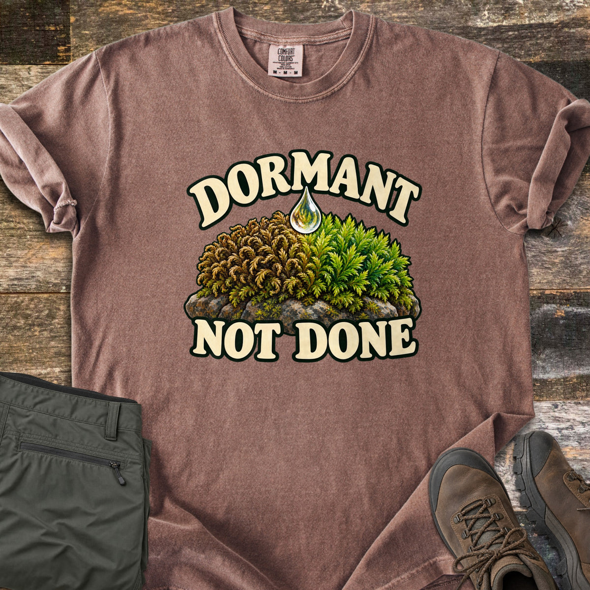 Dormant Not Done Comfort Colors T-shirt in Espresso, laid flat. Dormant arches in cream letters over moss on a rock, half dry and brown, half bright green, with a water drop above and Not Done below. Folded olive pants and brown hiking boots sit on rustic wood planks.