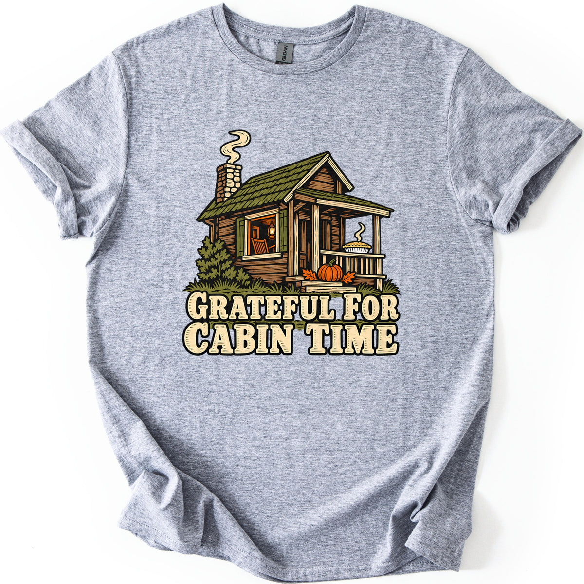 Grateful For Cabin Time Comfort Colors T-shirt in Sport Grey, laid flat. A log cabin with a green shingled roof, a smoking stone chimney, a steaming pie on the porch rail and a pumpkin by the steps sits above the words Grateful For Cabin Time. Sleeves rolled, on white.