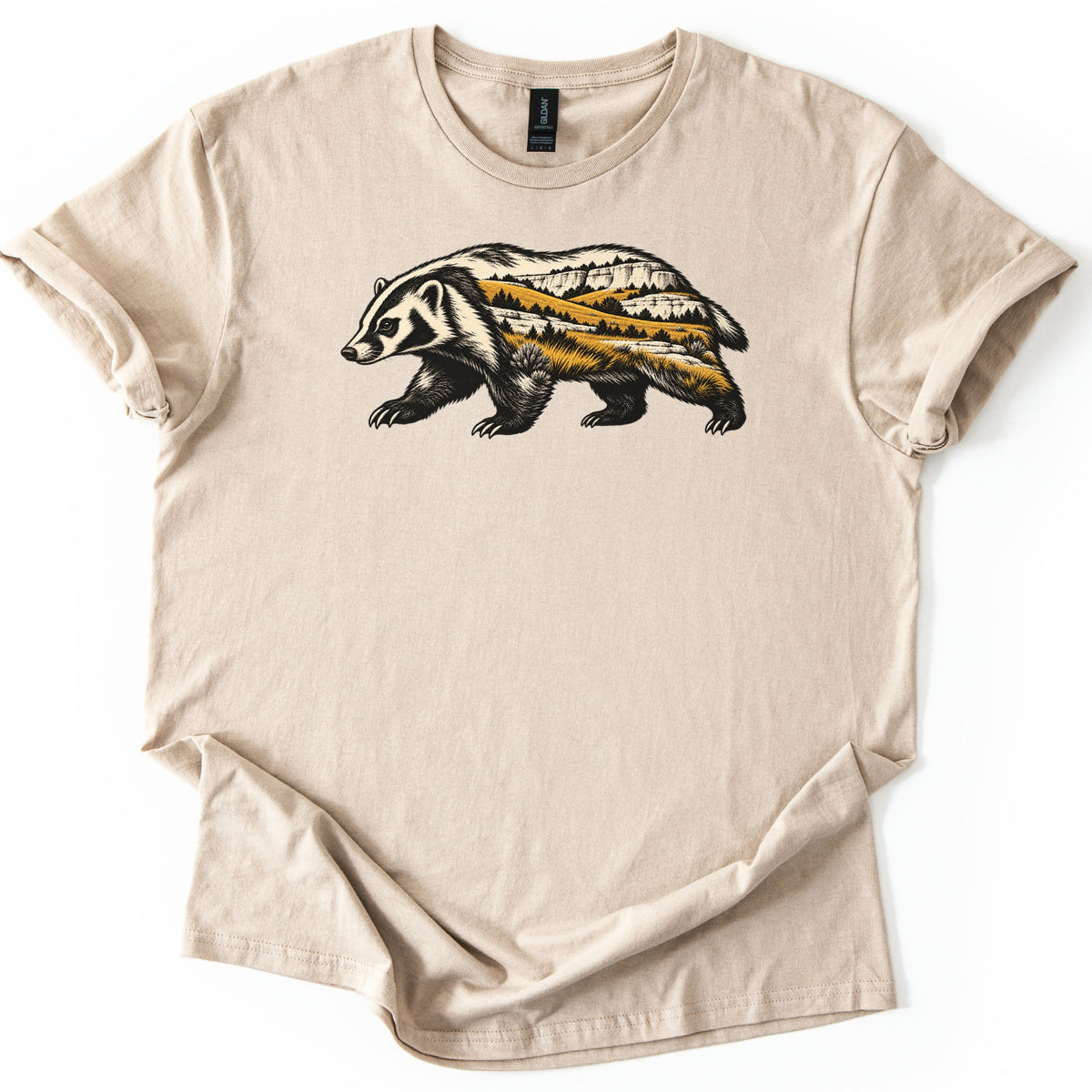 Badger Prairie Bluffs Comfort Colors T-shirt in Sand, laid flat. A badger walking left, its body filled with a scene of white bluffs, golden prairie grass and dark pine trees, with no words. The shirt lies on a white surface with cuffed sleeves and a loosely folded hem.