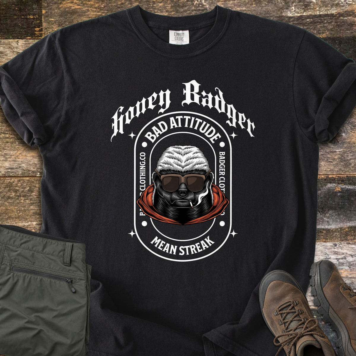 Bad Vibes Badger Comfort Colors T-shirt in Black, laid flat. A honey badger in sunglasses and a red hood smokes a cigarette in an arch reading Bad Attitude, Badger Clothing.co, Mean Streak, under Honey Badger in white gothic text. Olive pants and brown boots lie on wood.