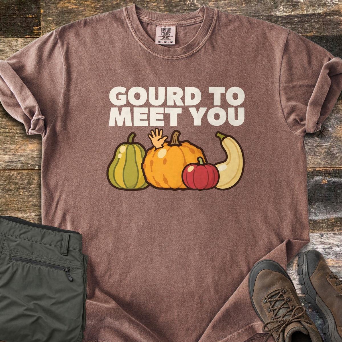 Gourd To Meet You Comfort Colors T-shirt in Espresso, laid flat. Off-white words Gourd To Meet You above cartoon gourds: a green striped squash, an orange pumpkin with a waving hand, a red pumpkin and a cream squash. Olive pants and brown hiking boots sit on wood planks.
