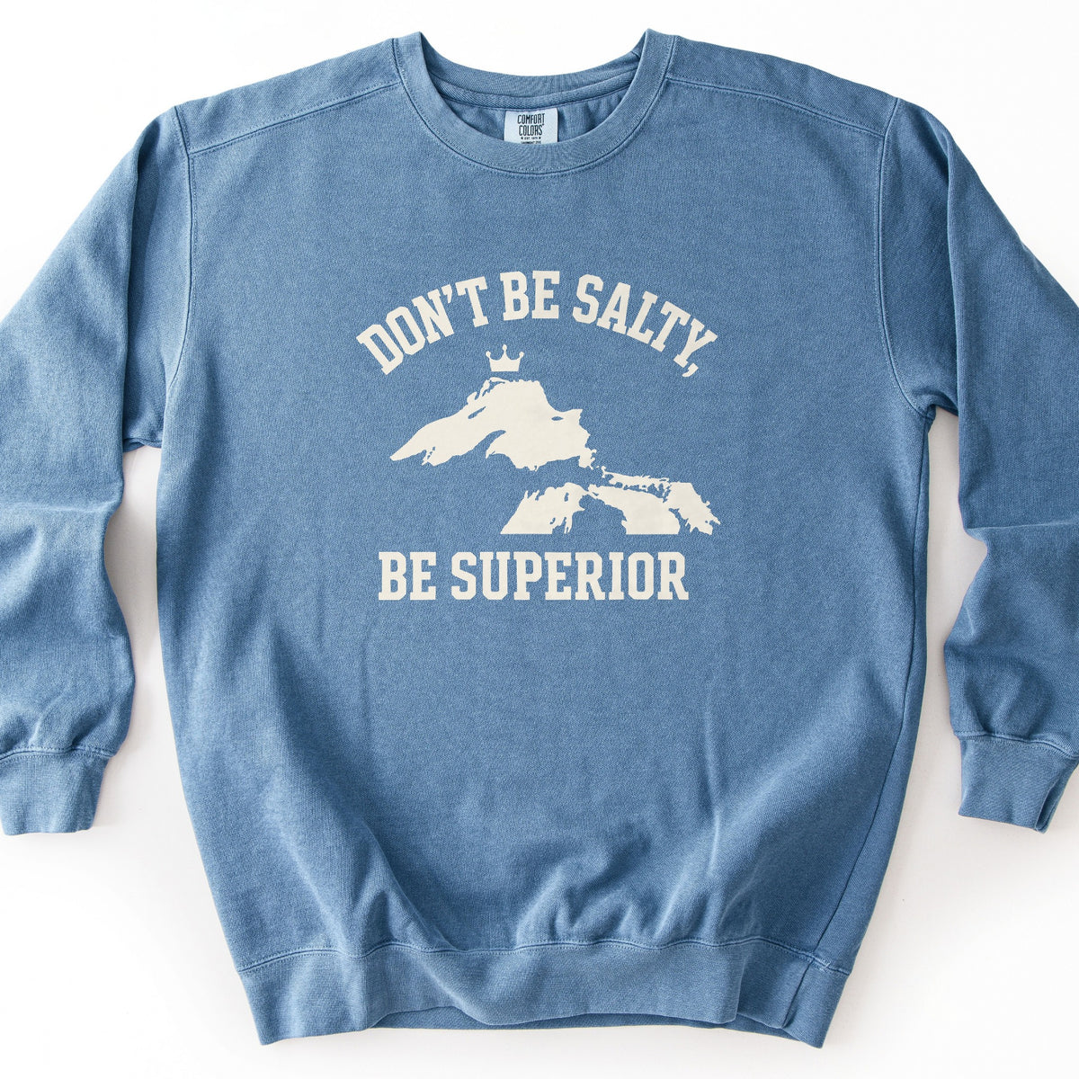 Be Superior Comfort Colors crewneck sweatshirt in Blue Jean, laid flat. Cream print of the words Don't Be Salty, arched above a silhouette of Lake Superior topped with a small crown, with Be Superior below. Laid on a white background.