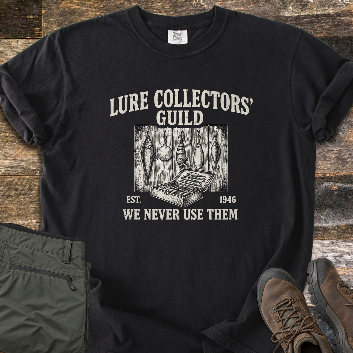 Lure Collectors Guild Comfort Colors T-shirt in Black, laid flat. A vintage-style drawing of five fishing lures hanging on wood planks above an open tackle box, framed by Lure Collectors' Guild, Est. 1946, We Never Use Them. Olive pants and brown hiking boots sit on wood.