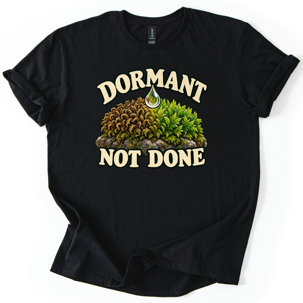 Dormant Not Done Comfort Colors T-shirt in Black, laid flat. Cream letters read Dormant above and Not Done below a clump of moss on a rock, half dry and brown, half lush green, with a water drop above it. Shown on a white surface with cuffed sleeves.