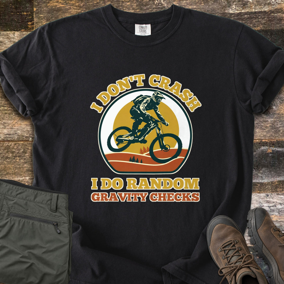 I Don't Crash I Do Random Gravity Checks Comfort Colors T-shirt in Black, laid flat. A mountain biker in a full-face helmet rides over orange hills and pines before a yellow sun, between the words I Don't Crash and I Do Random Gravity Checks. Olive pants and brown hiking boots lie on weathered wood.