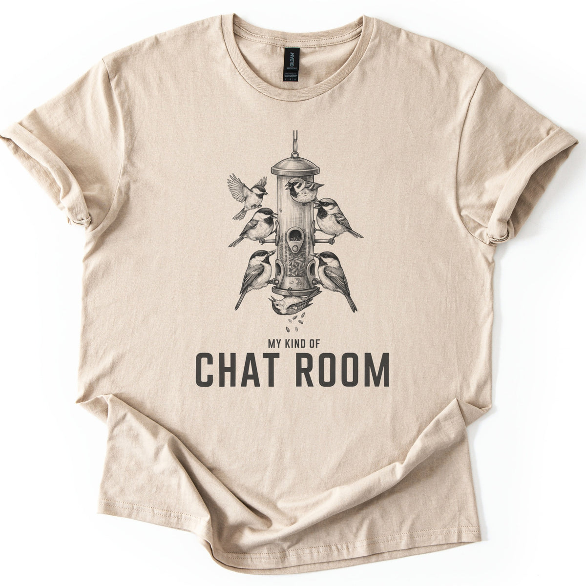 My Kind Of Chat Room Comfort Colors T-shirt in Sand, laid flat. A charcoal sketch of chickadees and sparrows perched around a hanging tube bird feeder, one upside down below spilling seeds, above the words My Kind Of Chat Room. Sleeves rolled, on a white background.