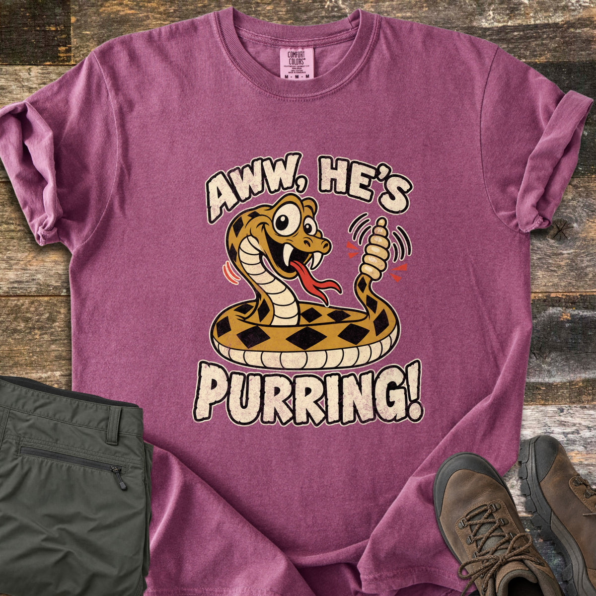 Aww He's Purring Comfort Colors T-shirt in Berry, laid flat. A coiled cartoon rattlesnake with big eyes, fangs and a red forked tongue rattles its tail under Aww, He's and above Purring! Olive hiking pants and brown hiking boots lie nearby on rustic wood boards.