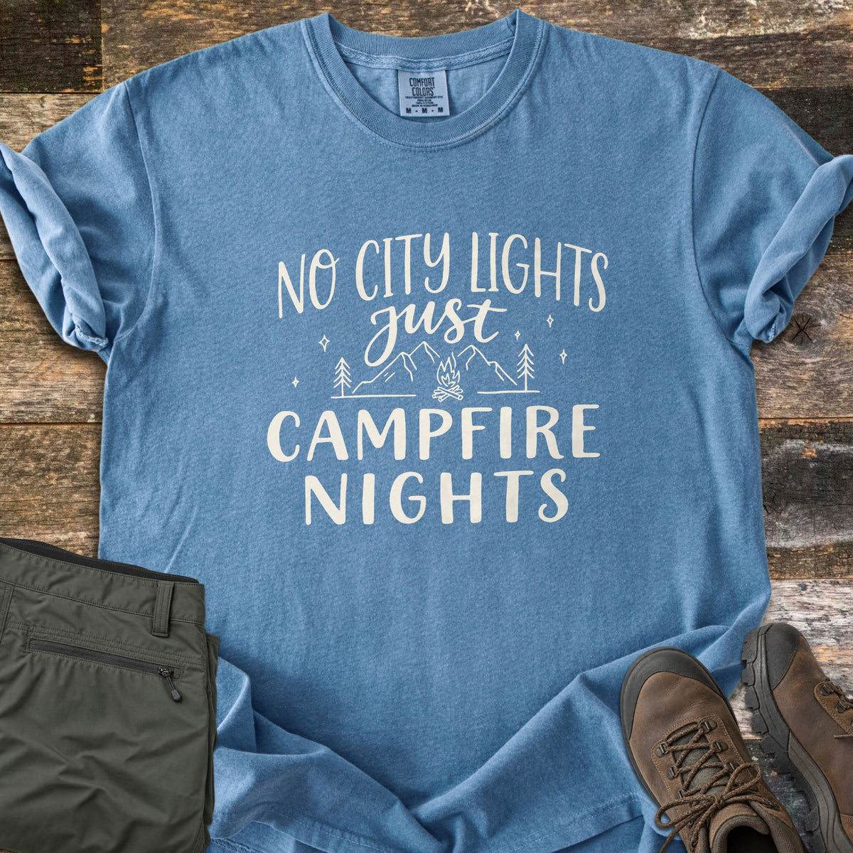 Campfire Nights Comfort Colors T-shirt in Blue Jean, laid flat. The words No City Lights just Campfire Nights in cream frame line art of mountains, pine trees, stars and a campfire. Laid on dark wood with olive hiking pants and brown hiking boots.