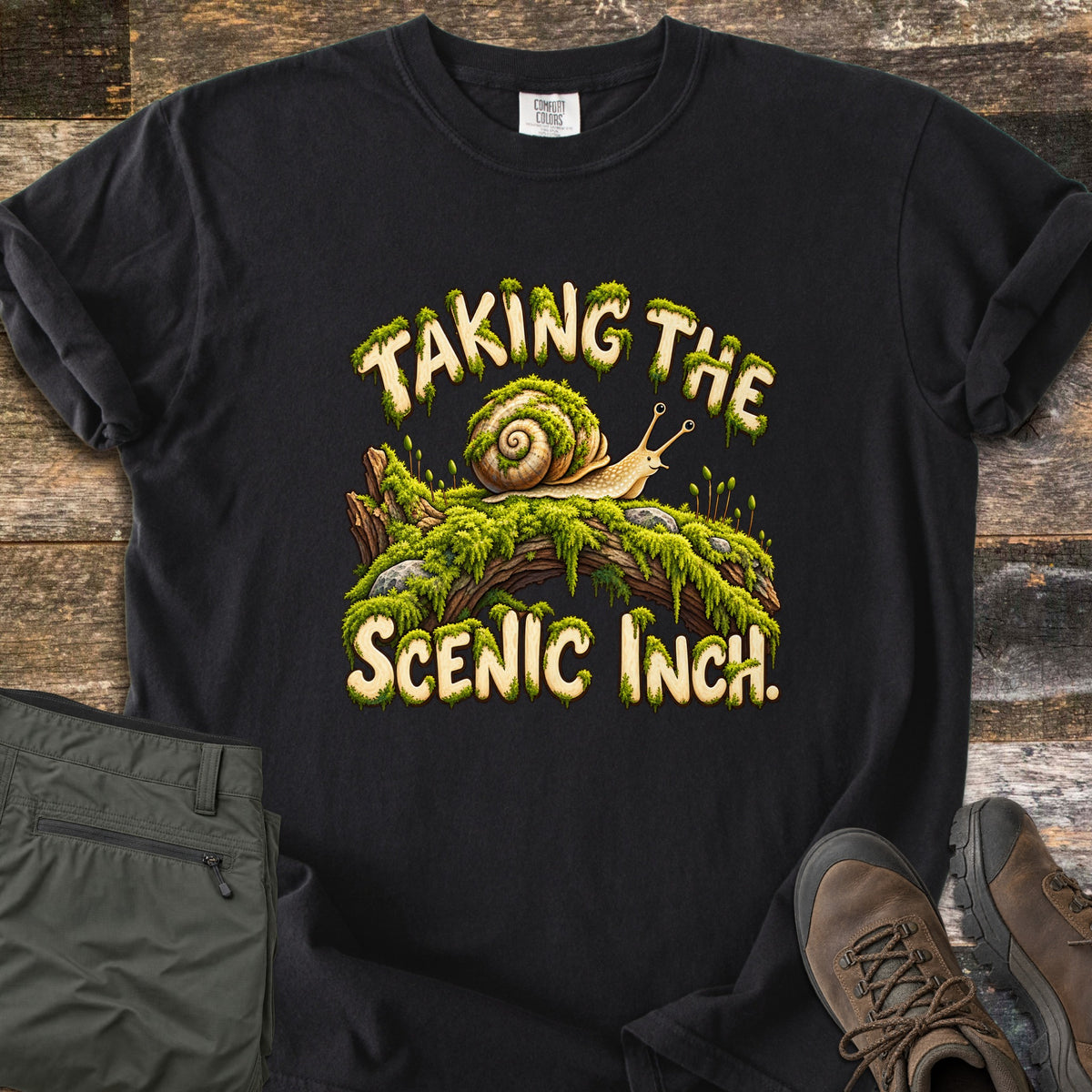 Taking the Scenic Inch Comfort Colors T-shirt in Black, laid flat. A smiling cartoon snail with a spiral shell crawls along a mossy log between the words Taking the Scenic Inch., lettered in cream with dripping moss. Olive hiking pants and brown boots sit on a weathered wood floor.