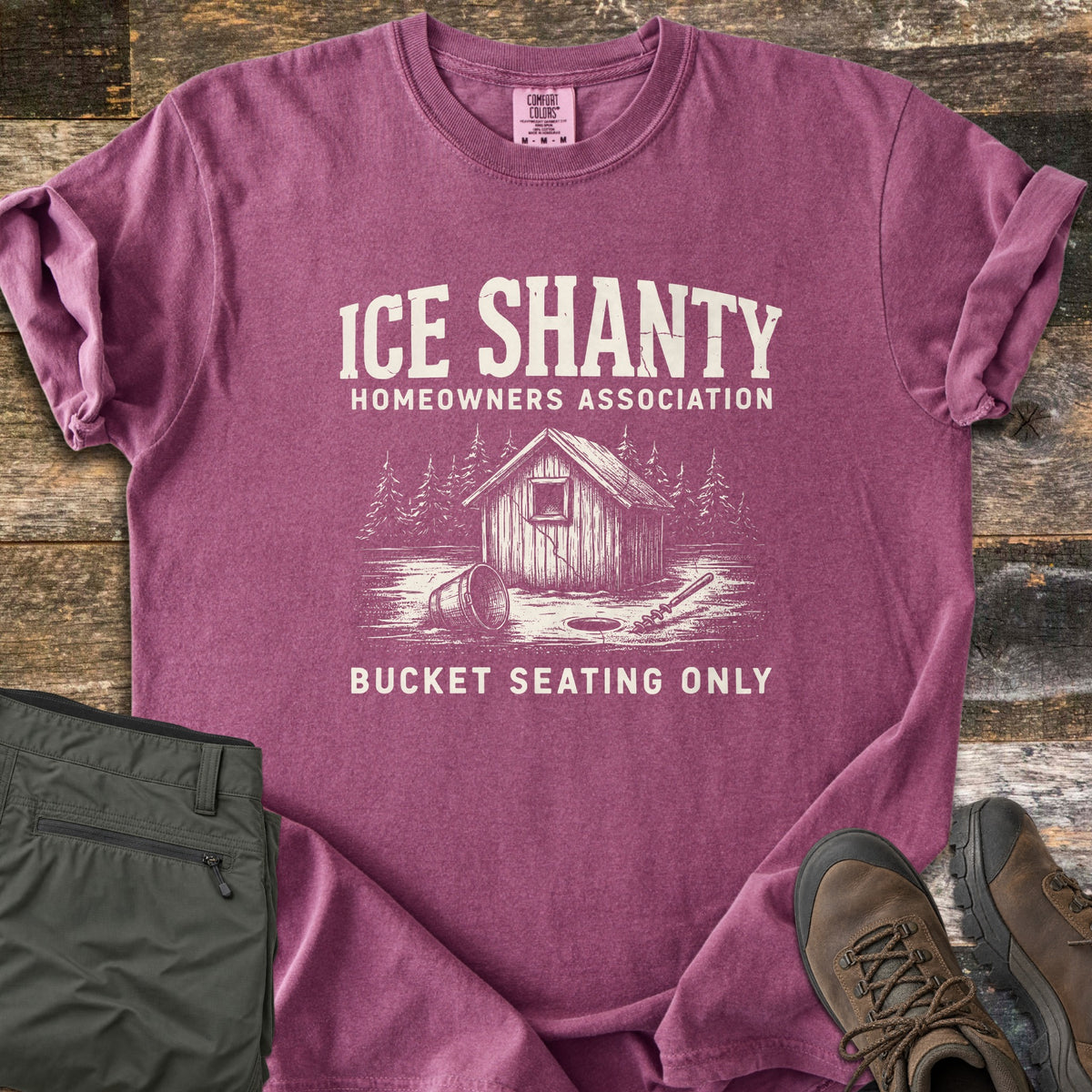 Ice Shanty Homeowners Association Comfort Colors T-shirt in Berry, laid flat. Cream print of a wooden ice shanty among pines, a tipped bucket, ice hole and auger, with the words Ice Shanty Homeowners Association and Bucket Seating Only. Olive pants and brown hiking boots sit on weathered wood.