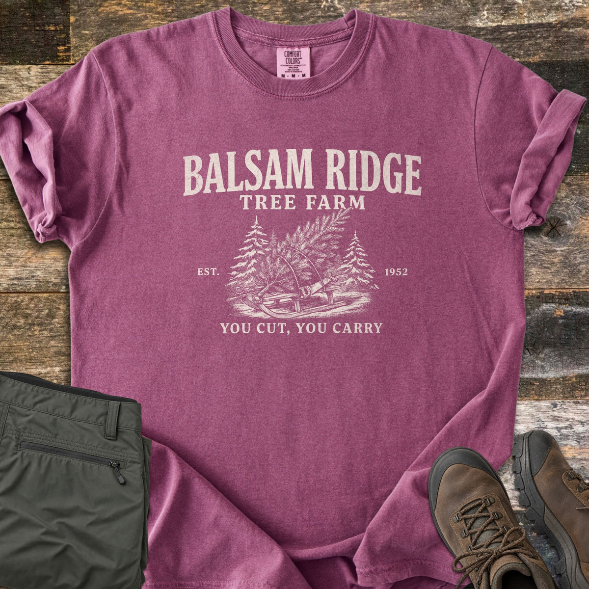 Balsam Ridge Tree Farm Comfort Colors T-shirt in Berry, laid flat. Cream print reads Balsam Ridge Tree Farm above a cut evergreen tied to a wooden sled with a bow saw between two pines, Est. 1952, and You Cut, You Carry. Olive pants and brown hiking boots sit on weathered wood.