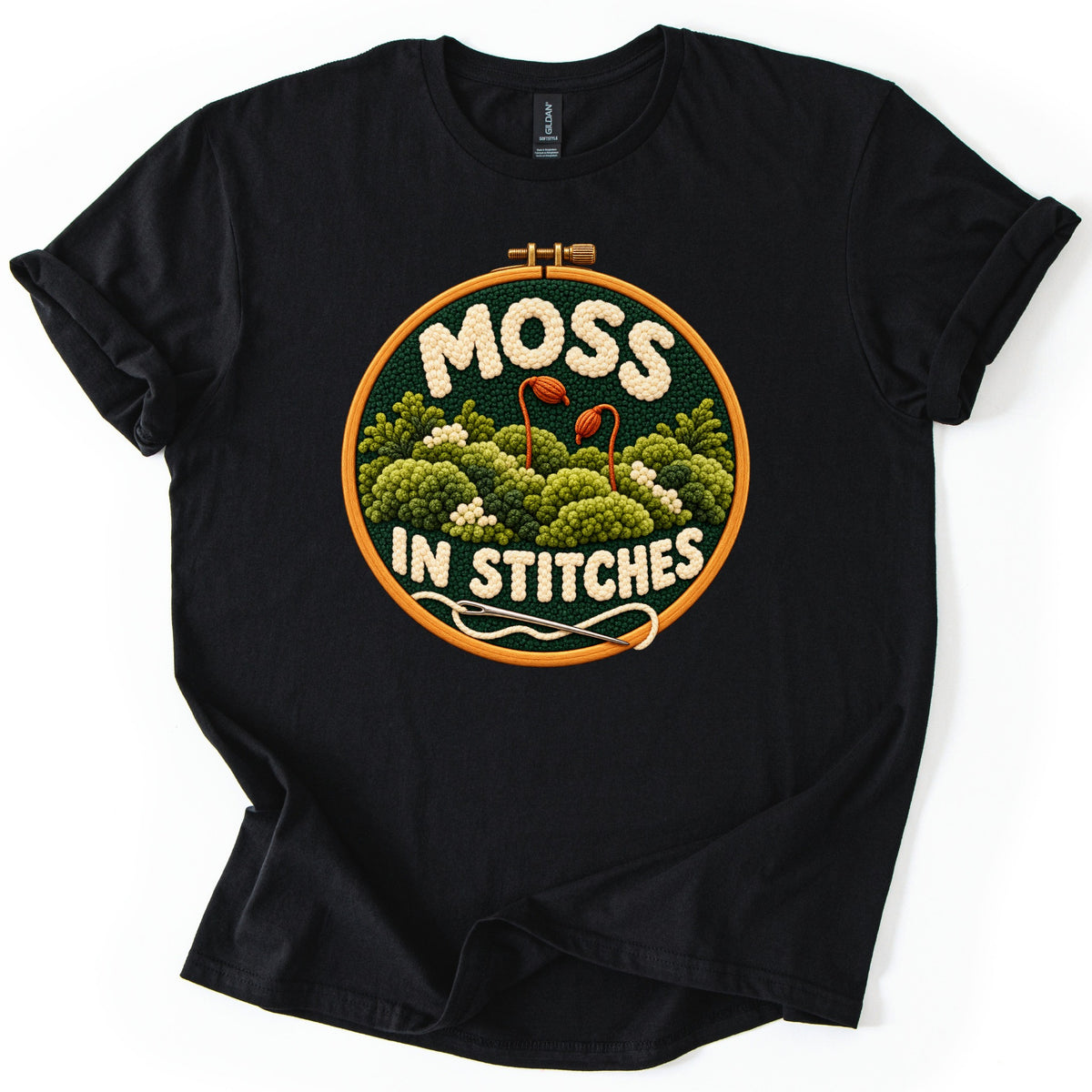 Moss in Stitches Comfort Colors T-shirt in Black, laid flat. An embroidery hoop shows textured green moss mounds and two orange spore stalks on dark green, with MOSS IN STITCHES in cream yarn and a threaded needle below. Sleeves rolled, on a white background.