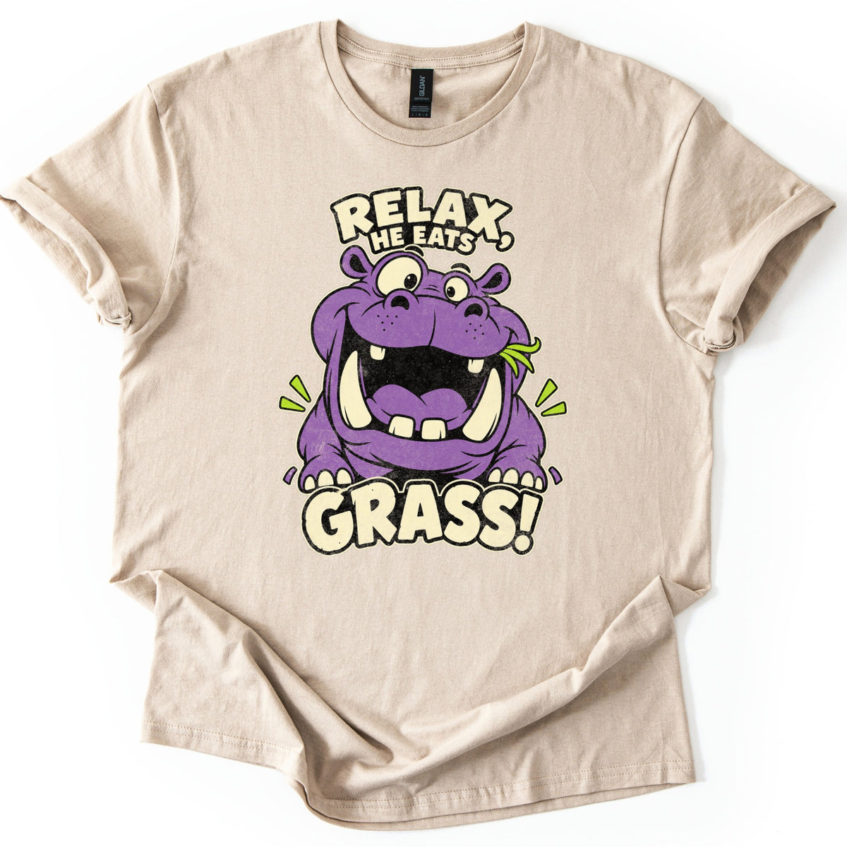 Relax He Eats Grass Comfort Colors T-shirt in Sand, laid flat. A grinning purple cartoon hippo with big tusks and a blade of grass in its mouth sits between the words RELAX, HE EATS and GRASS! in cream letters. The shirt lies on white with rolled sleeves.