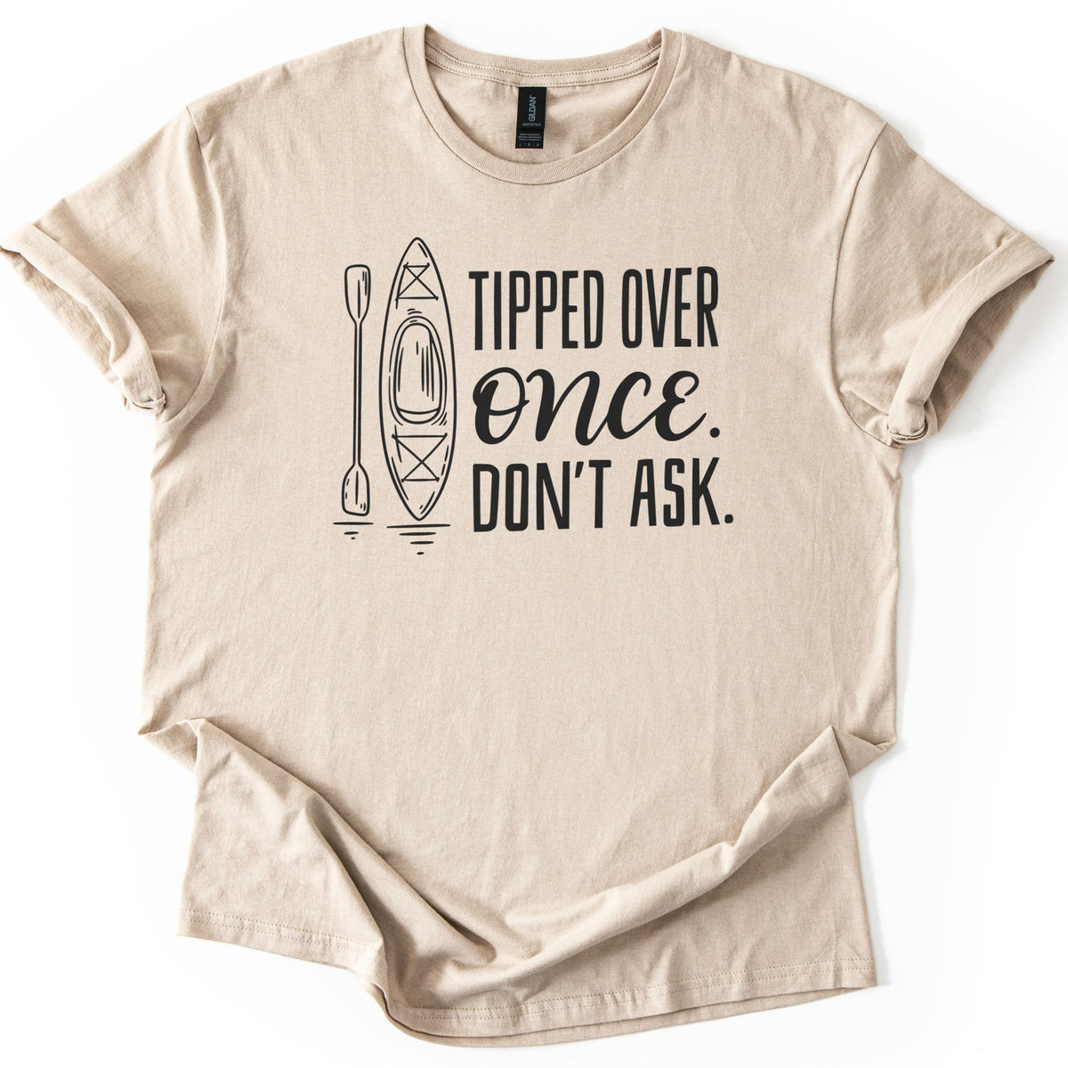 Tipped Over Once Kayak Comfort Colors T-shirt in Sand, laid flat. A black line drawing of an upright kayak and paddle with water ripples sits beside the words Tipped Over Once. Don't Ask., with Once in script. The shirt has cuffed sleeves on a white background.