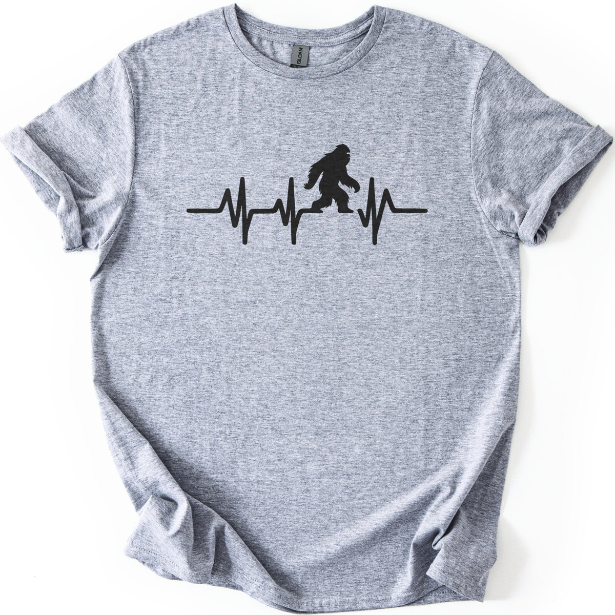 Yeti Heartbeat Comfort Colors T-shirt in Sport Grey, laid flat. A black heartbeat line runs across the chest, with a walking Bigfoot silhouette standing within it; there are no words. The sleeves are cuffed and the shirt lies on a white background.