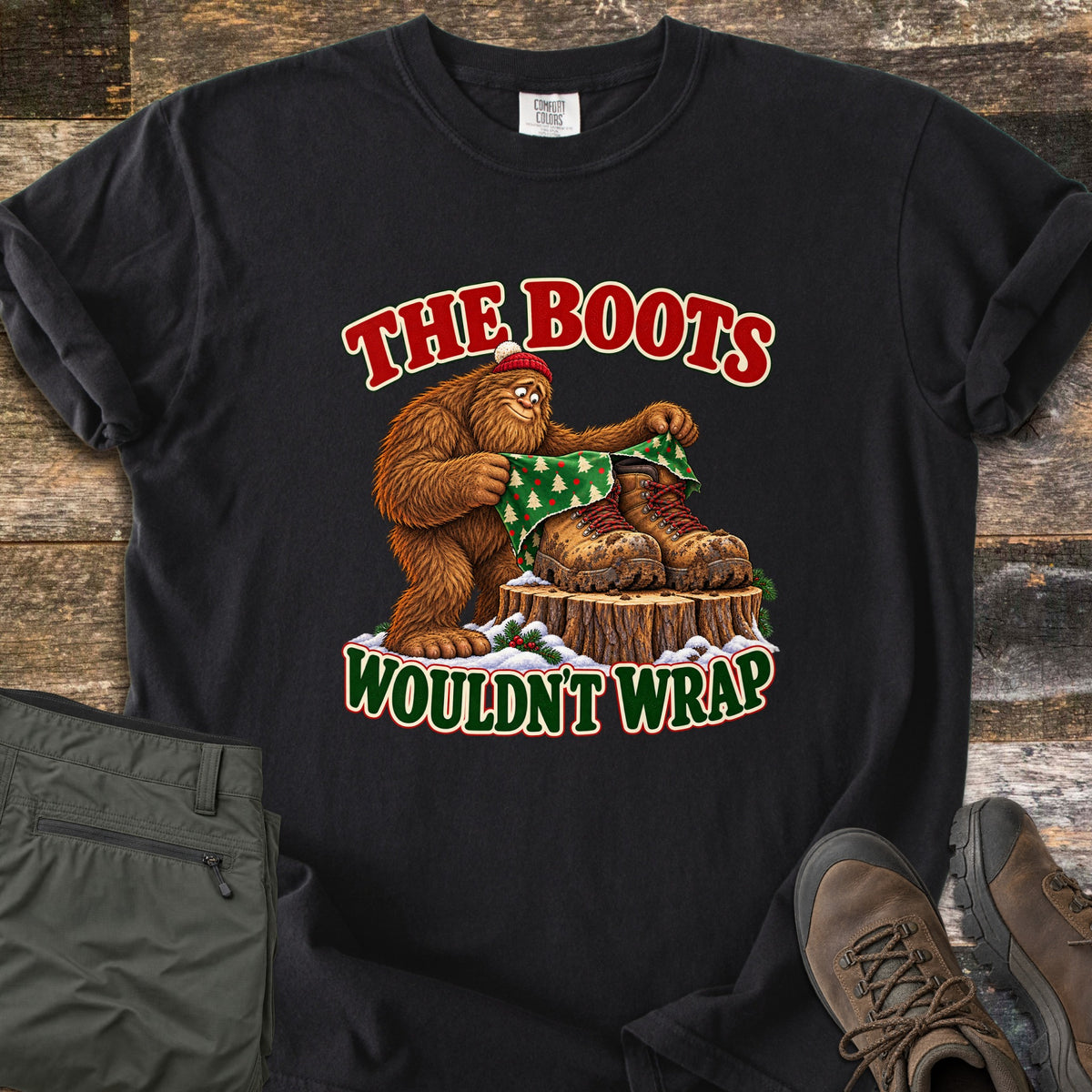 The Boots Wouldn't Wrap Comfort Colors T-shirt in Black, laid flat. A Bigfoot in a red beanie tries to wrap muddy hiking boots on a snowy stump in green Christmas tree paper, between the words The Boots Wouldn't Wrap. Olive pants and brown hiking boots lie beside it on weathered wood.