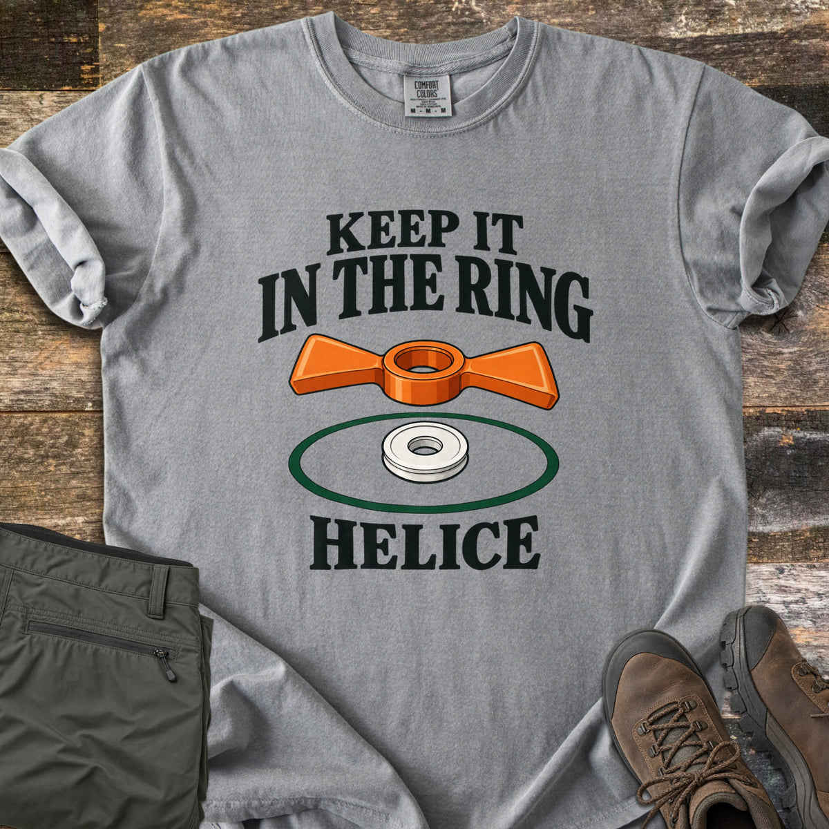 Keep It in the Ring Comfort Colors T-shirt in Grey, laid flat. An orange winged helice target floats above a white ring inside a green circle, between the words Keep It in the Ring and Helice. Olive pants and brown hiking boots sit beside it on weathered wood.