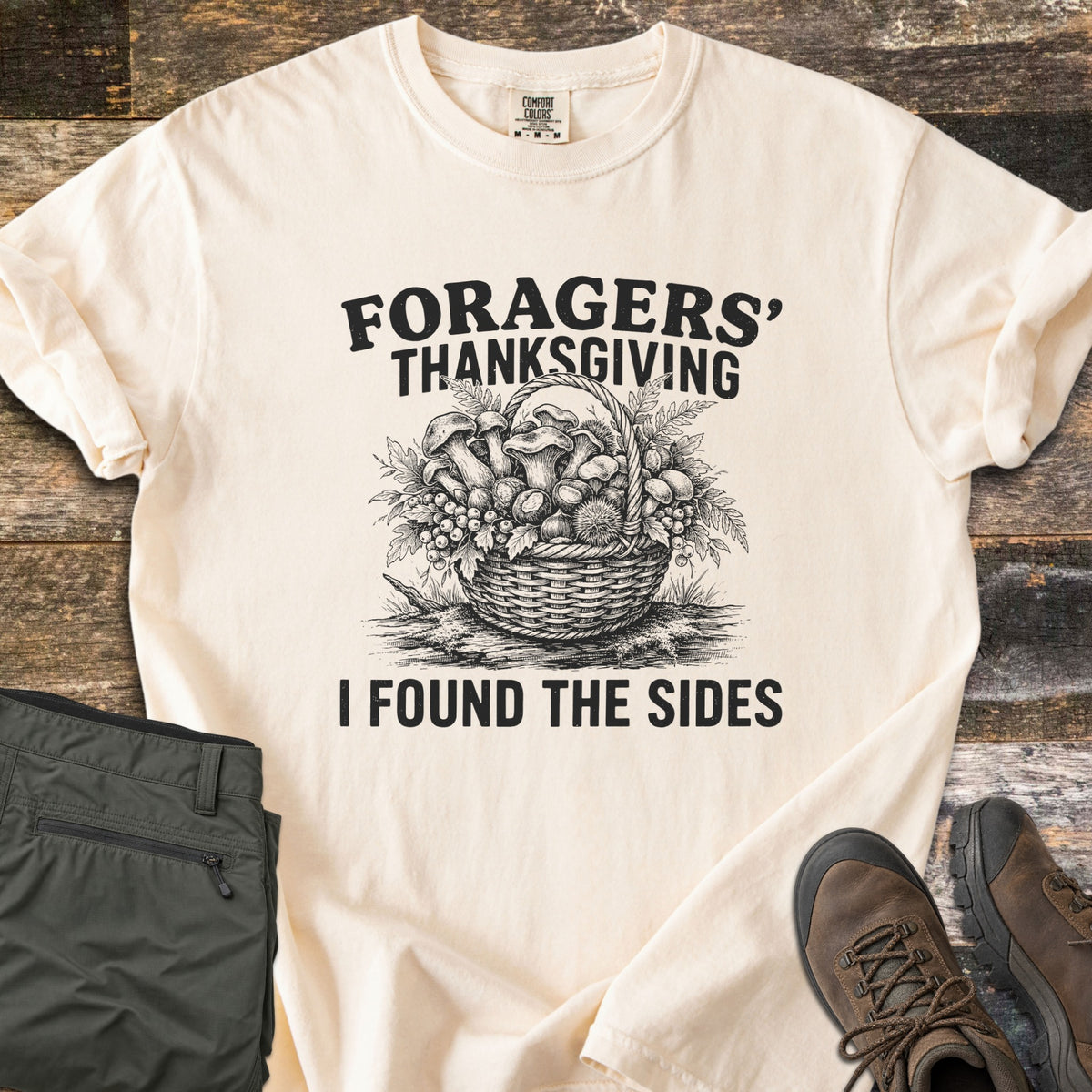 Foragers Thanksgiving Comfort Colors T-shirt in Ivory, laid flat. A black sketch of a woven basket full of mushrooms, berries and leaves sits between the words Foragers' Thanksgiving and I Found the Sides. Folded olive hiking pants and brown boots lie on weathered wood.