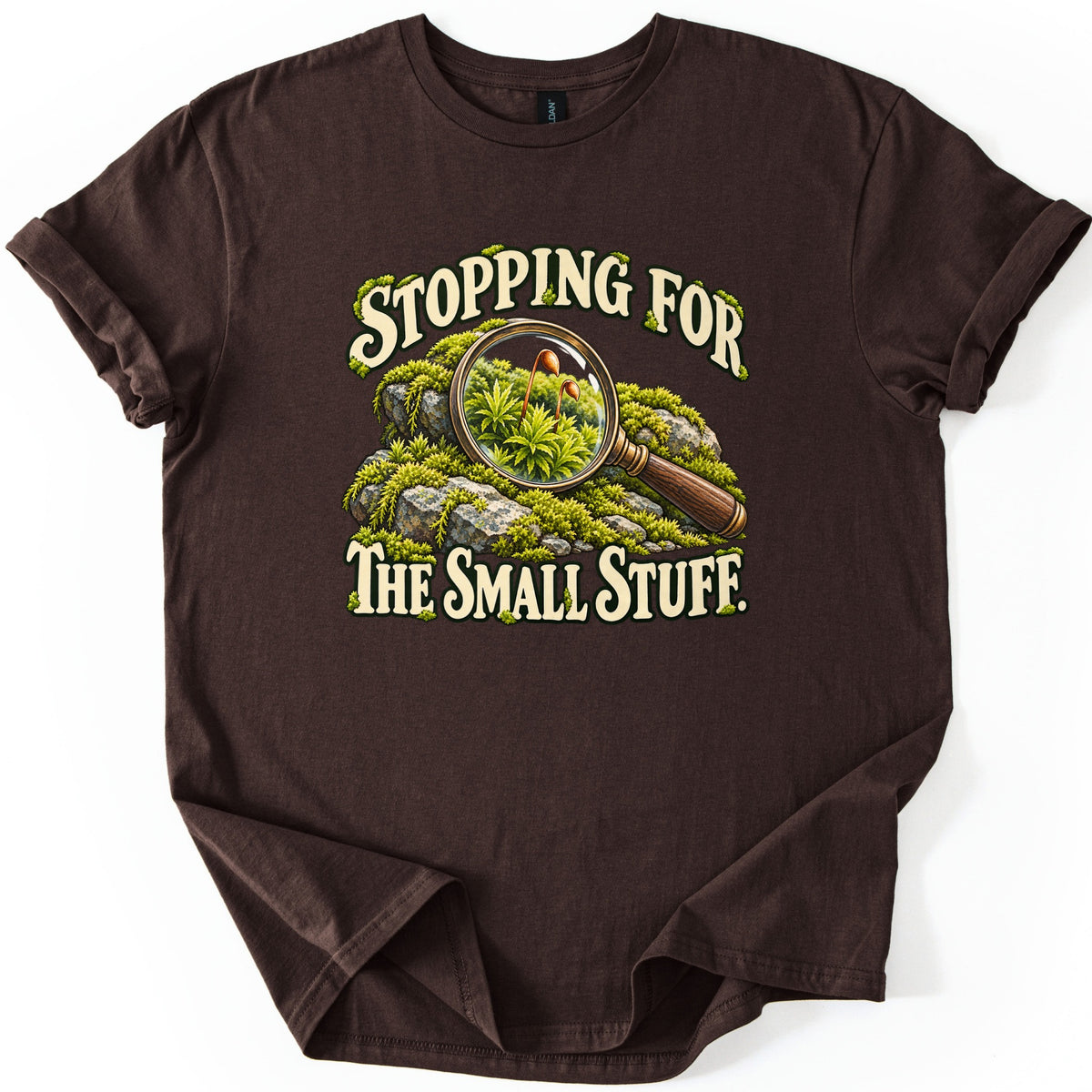 Stopping for the Small Stuff Comfort Colors T-shirt in Dark Chocolate, laid flat. A magnifying glass lies on moss-covered rocks, enlarging moss sprouts with tiny orange spore capsules, framed by the words Stopping For The Small Stuff. Sleeves are rolled on a white background.