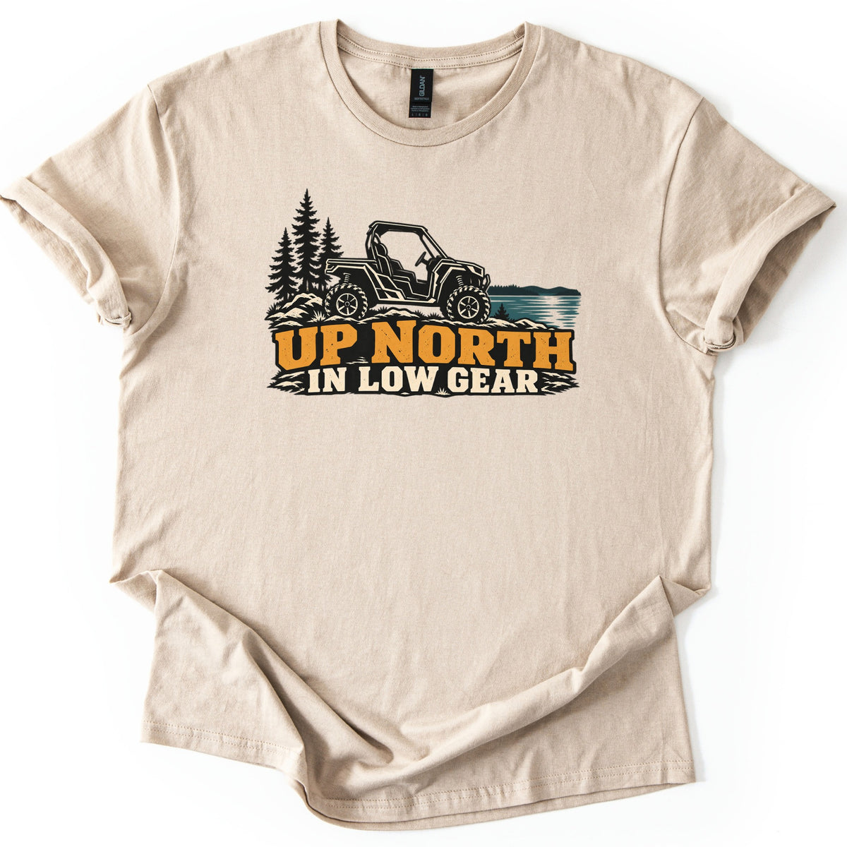 Up North in Low Gear Comfort Colors T-shirt in Sand, laid flat. A black side-by-side UTV on rocky ground between pine trees and a blue lake sits above the words UP NORTH in orange and IN LOW GEAR in cream. The shirt lies on a white surface with cuffed sleeves.