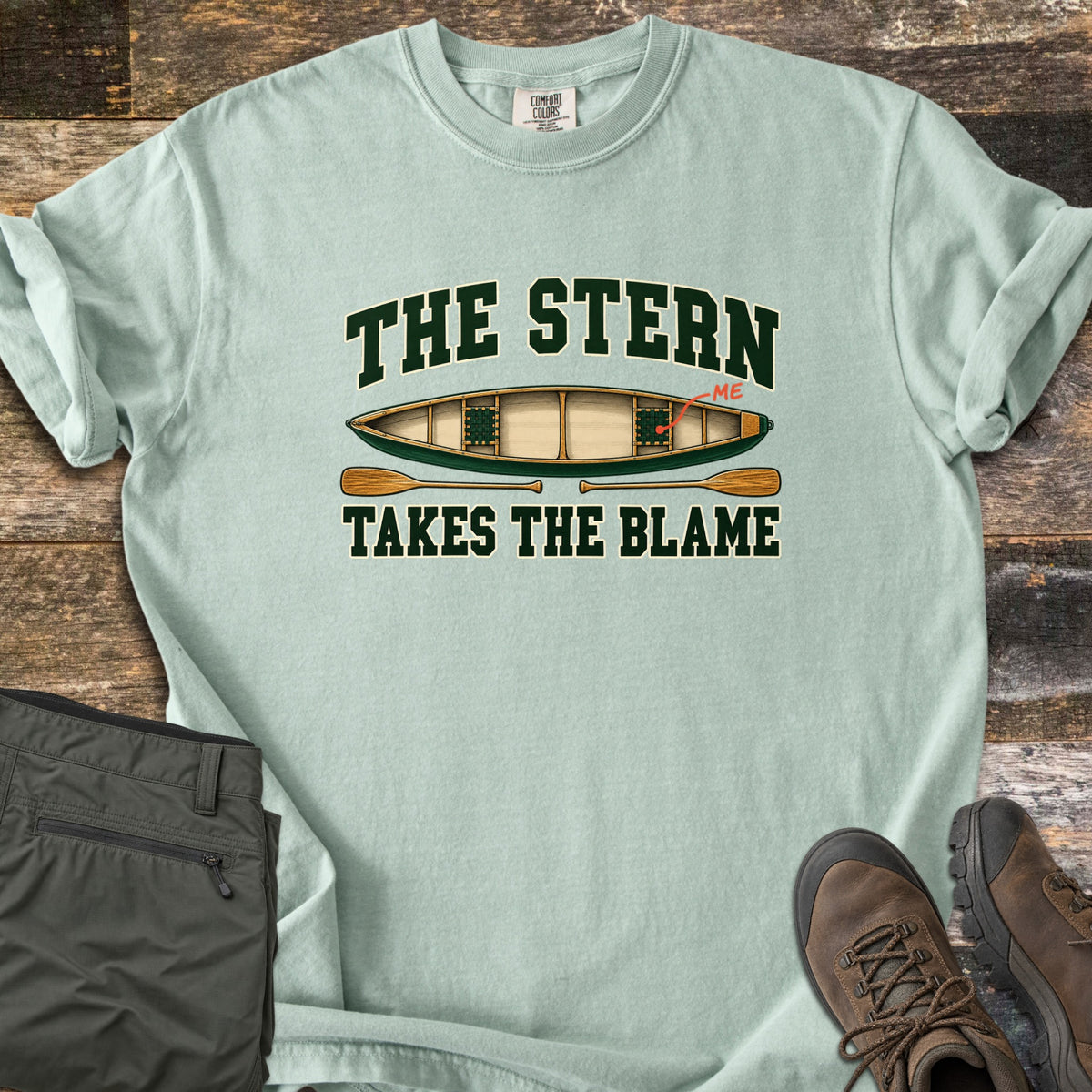The Stern Takes the Blame Comfort Colors T-shirt in Bay, laid flat. A green canoe seen from above, with a red arrow pointing to the back seat labeled ME, sits under The Stern and over two wooden paddles and Takes the Blame. Olive hiking pants and brown boots rest on rustic wood.